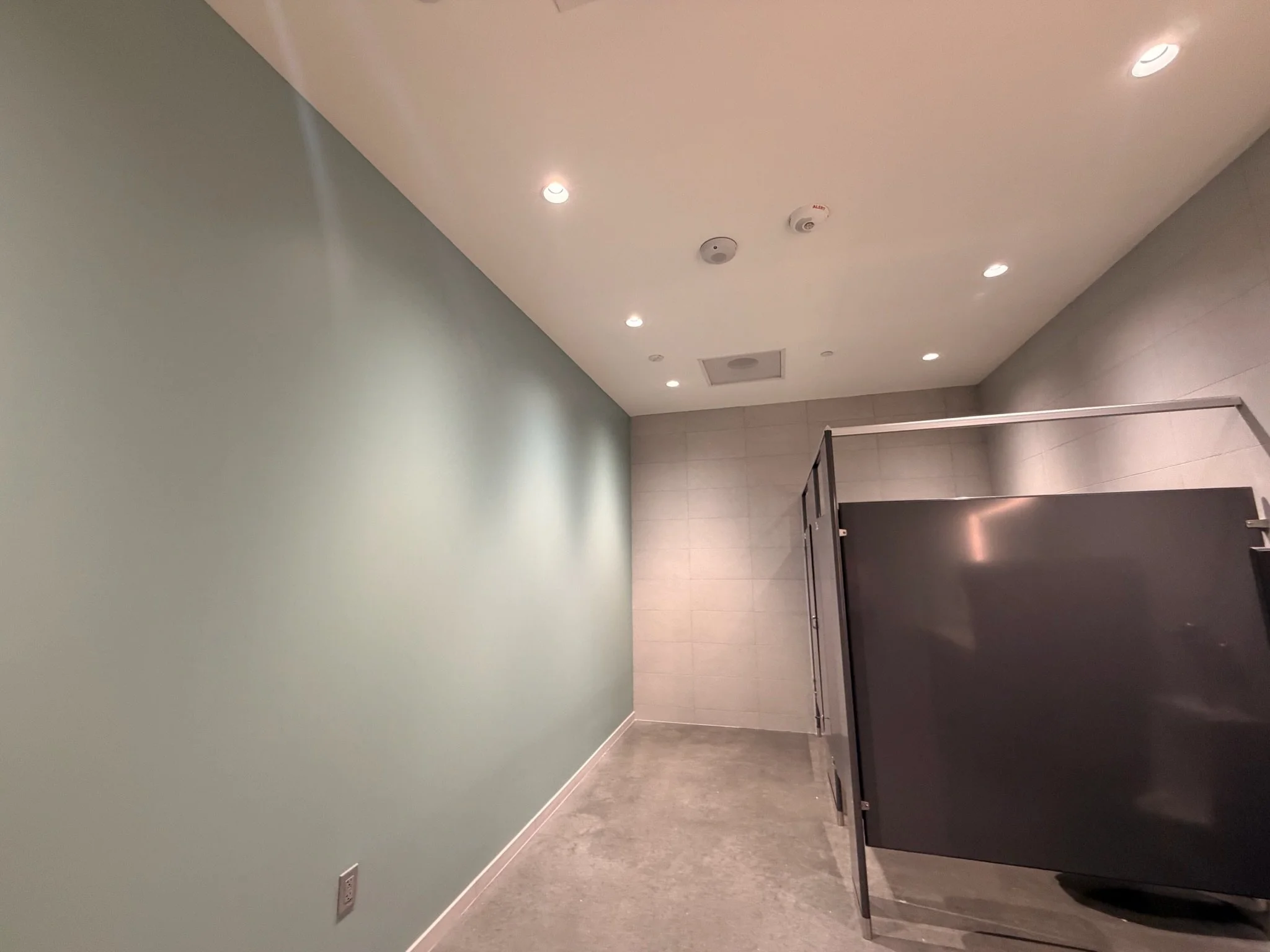 A restroom with mint green walls, beige floor tiles, and a ceiling with recessed lighting and smoke detectors. There is a partial black stall door on the right side.