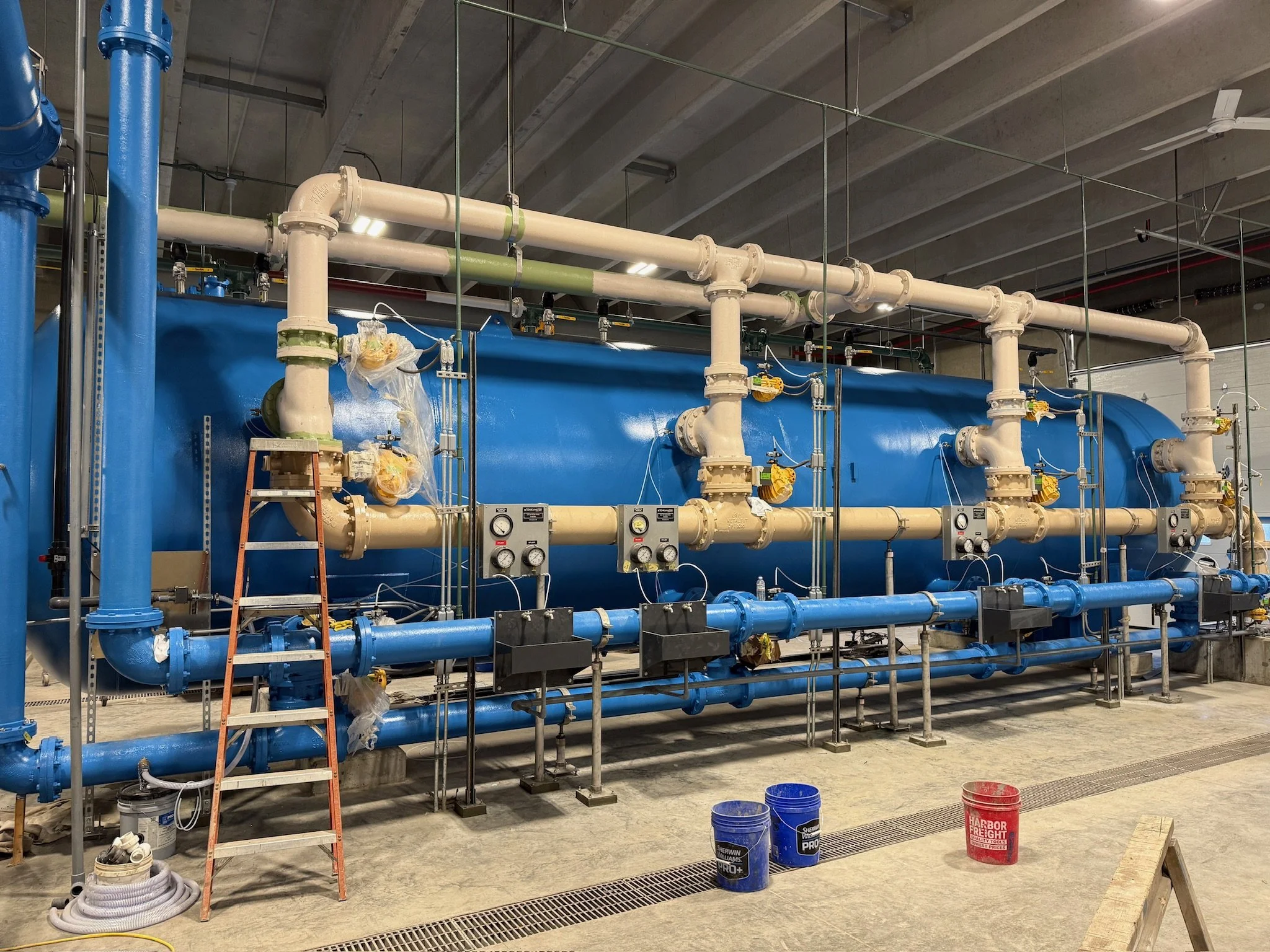 Industrial plumbing setup with large blue tank, white pipes, valves, gauges, and construction ladders in a warehouse or factory setting.