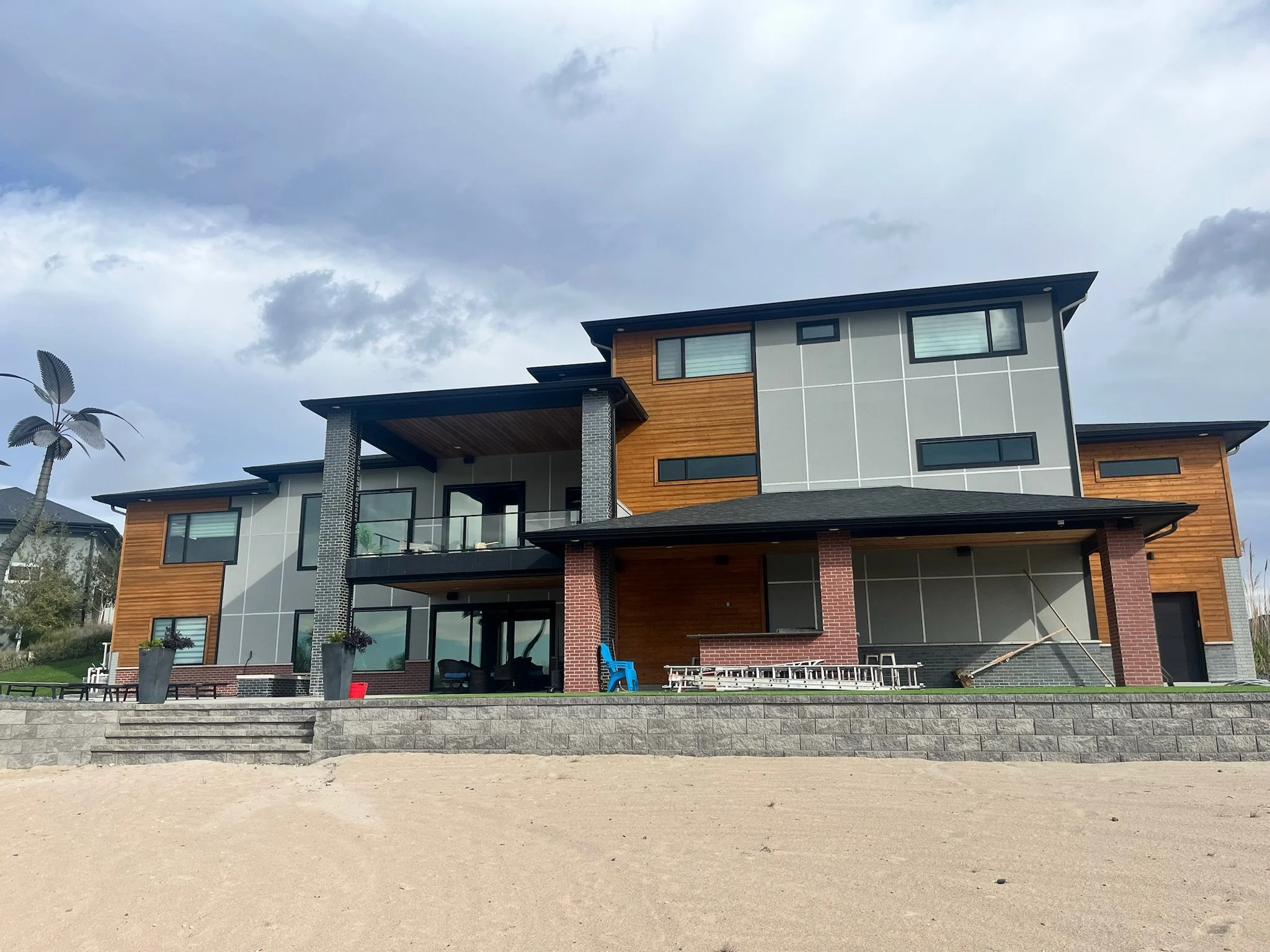 A modern multi-story house with wooden accents, large windows, and a balcony, situated in a suburban or semi-rural area with a cloudy sky overhead.