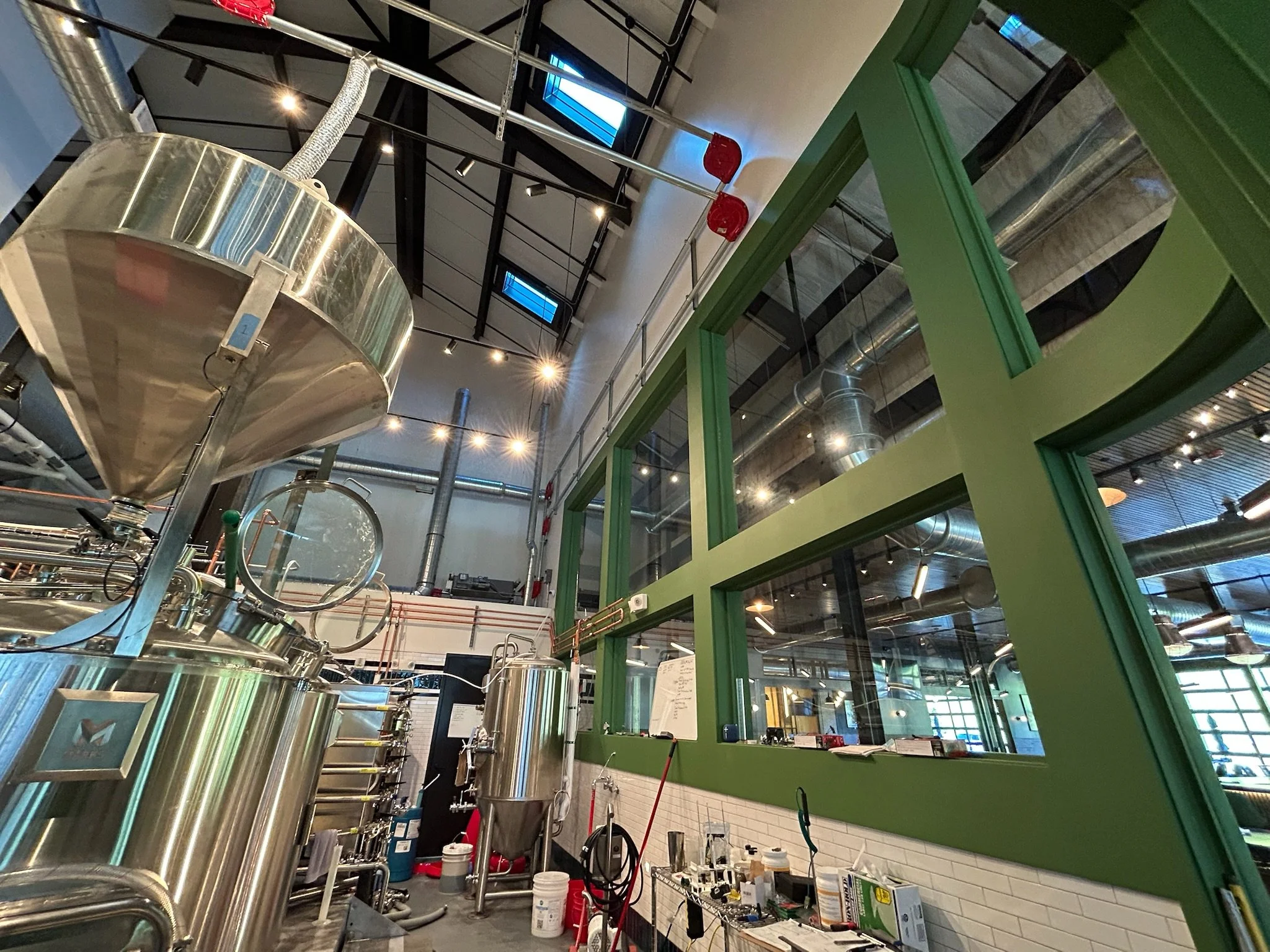 Industrial kitchen or brewery with large stainless steel tanks, green framing, and equipment on counters. Exposed ductwork and high ceiling with industrial lighting.