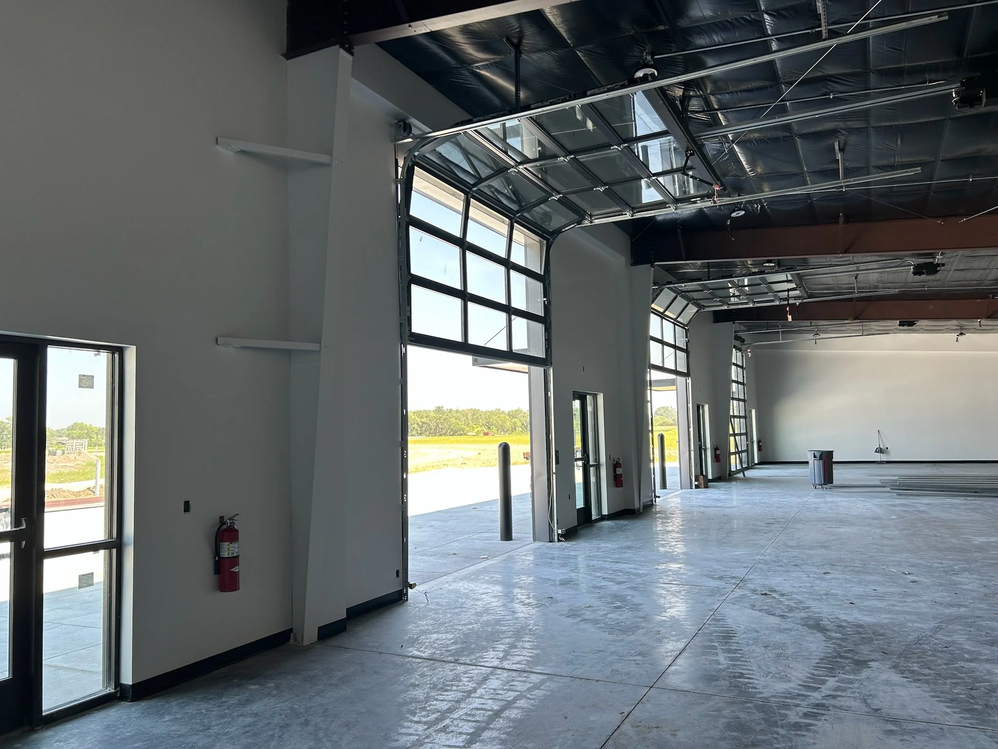 Interior of a building with large glass garage doors, high black ceiling, and natural light coming in. There are some safety fire extinguishers on the wall and light fixtures hanging from the ceiling.