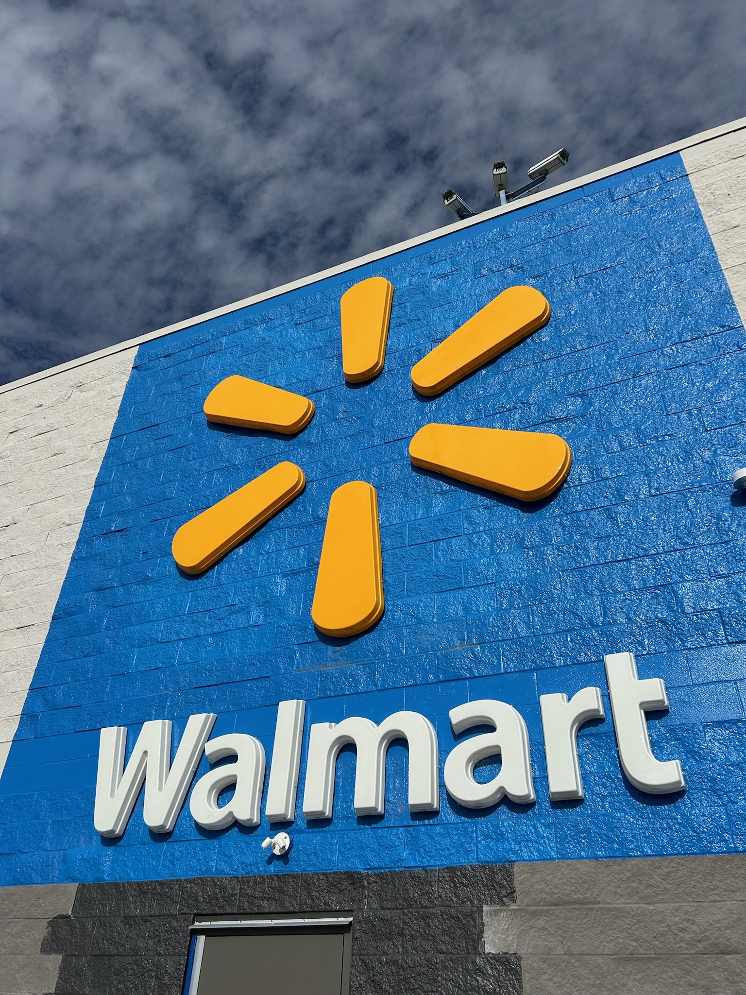 Close-up view of a Walmart store sign with the Walmart logo and part of the store's exterior wall, showing surveillance cameras on the top corner.