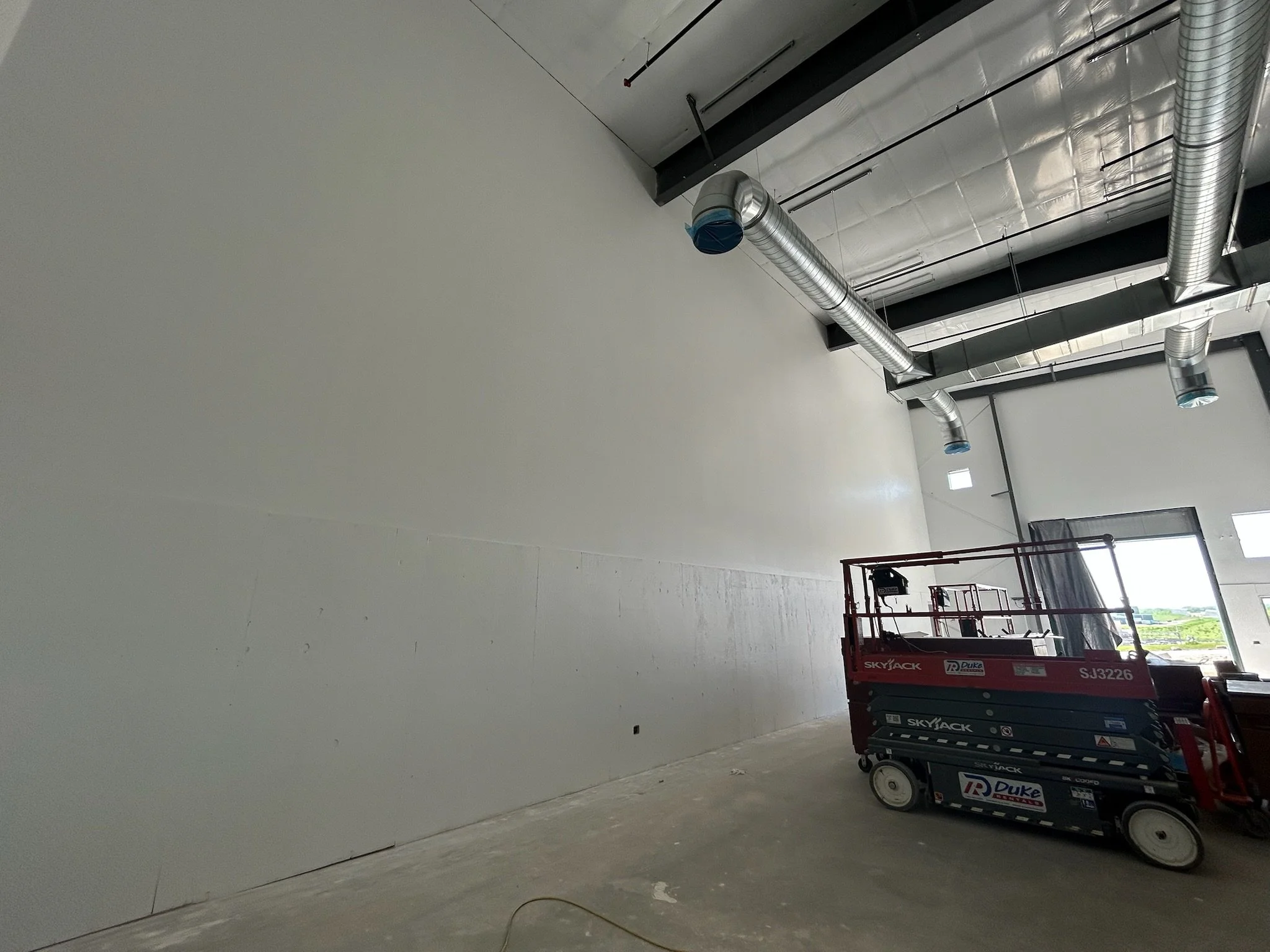 Inside a large industrial building under construction with exposed ductwork on the ceiling, a red scissor lift parked on a concrete floor, and a large blank white wall.