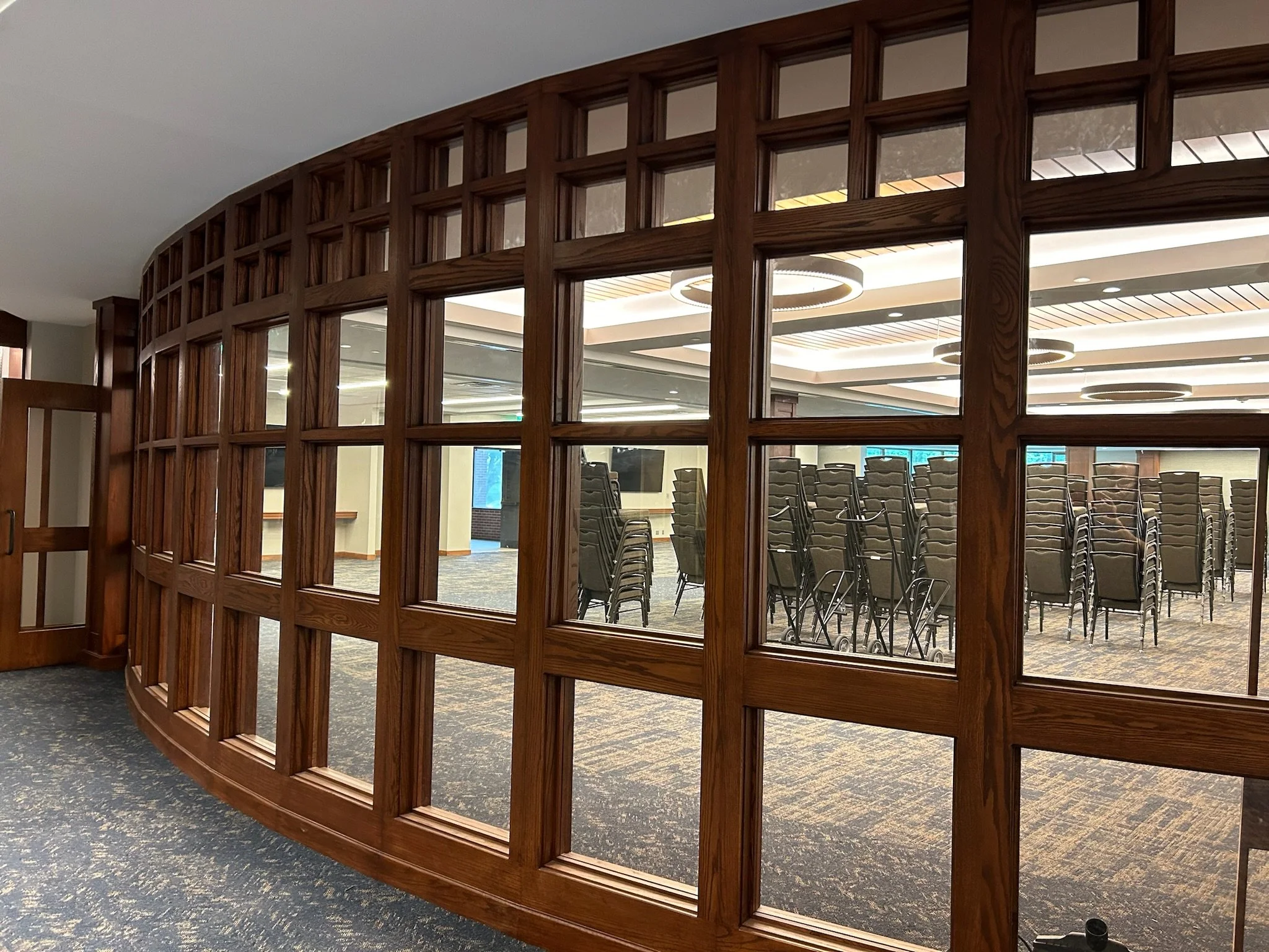 A large wooden lattice partition separates a conference room from the rest of the space. Inside the room, chairs are stacked and arranged in rows, with multiple circular ceiling lights overhead. The room has windows and bright lighting.