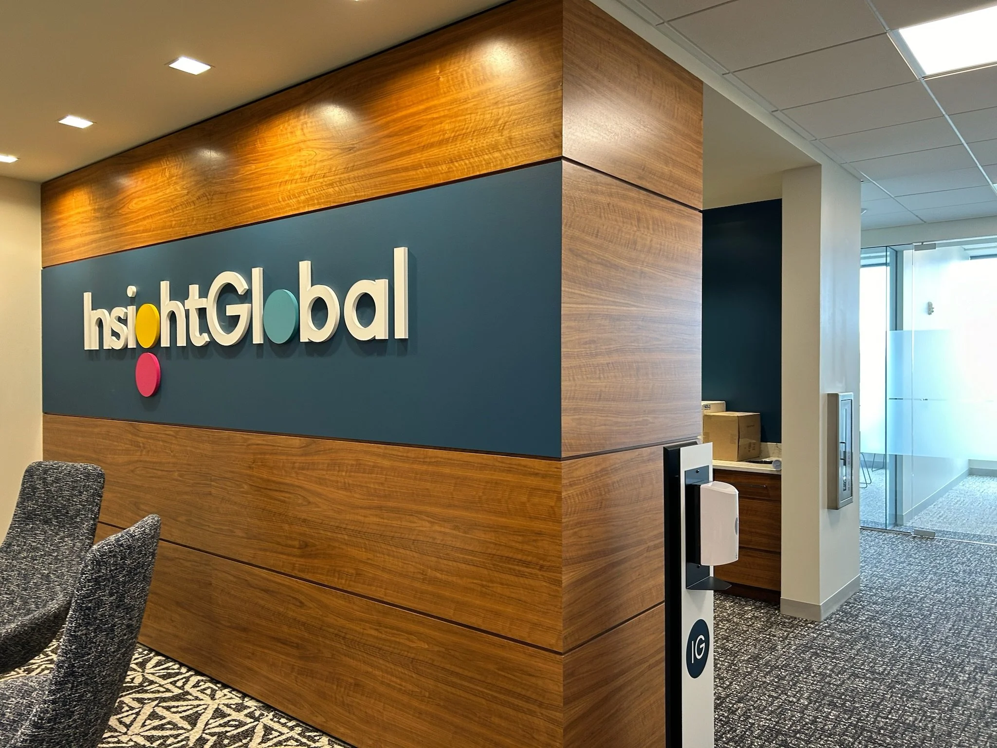 Interior of a modern office with a wood-paneled wall featuring the 'Insight Global' logo in white, colorful dots, and a blue background. There are black and gray patterned chairs and a window with open blinds allowing natural light.