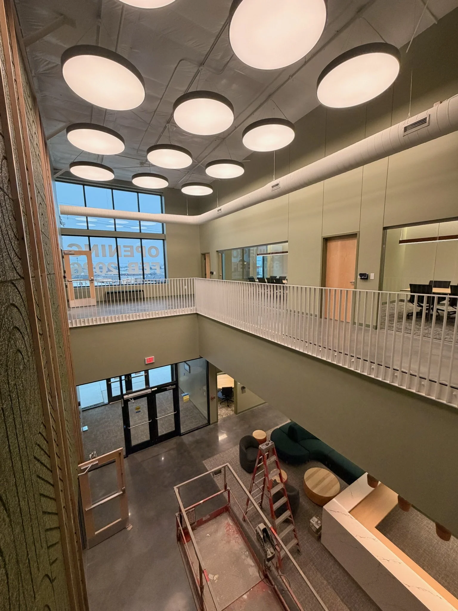 Interior of a modern building with high ceiling lights, a second-floor balcony with glass walls, and a spacious lobby area with seating and a ladder.