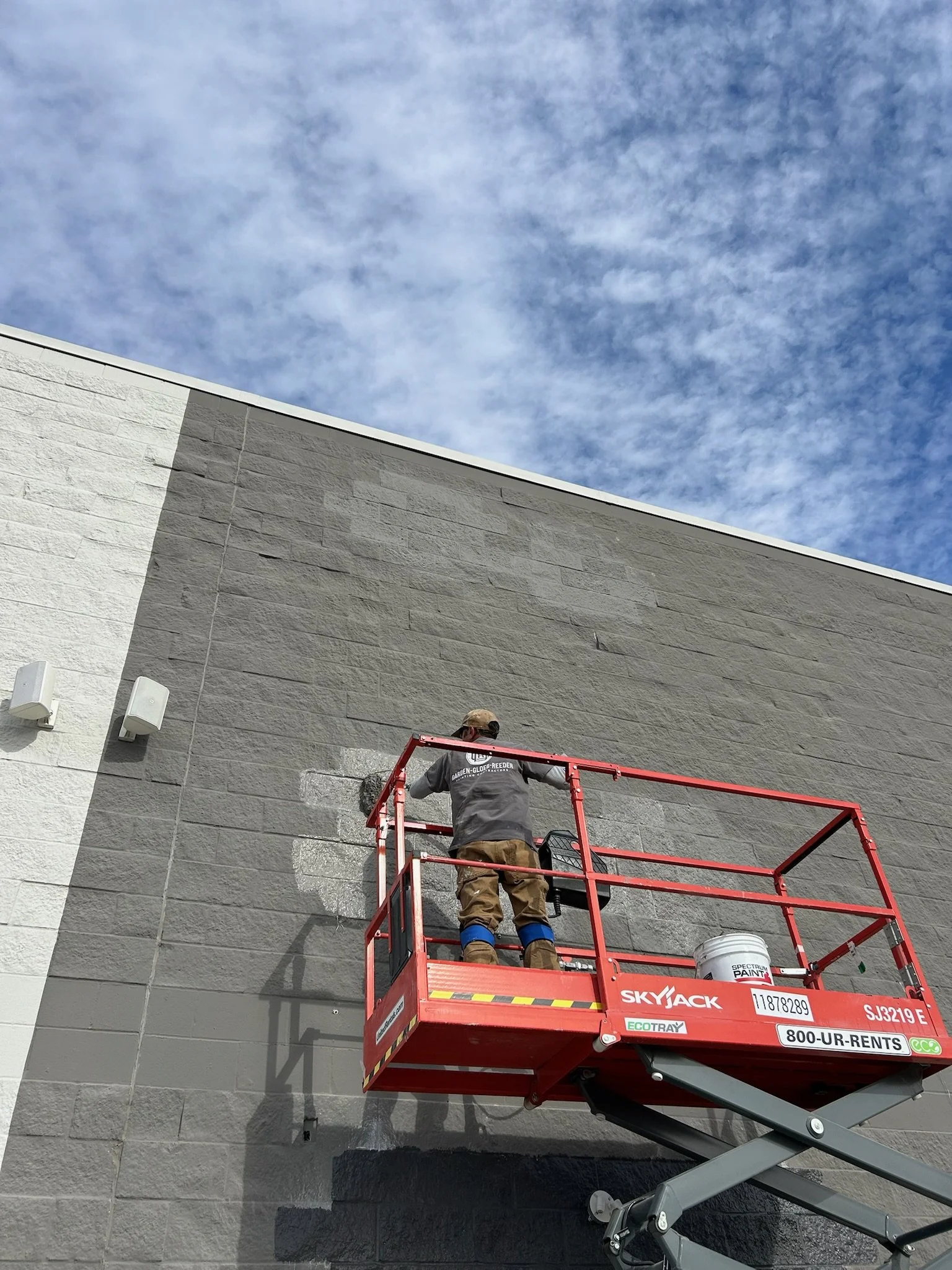 A worker on a red lift applying or repairing paint on a gray brick wall outside under a blue sky with scattered clouds.