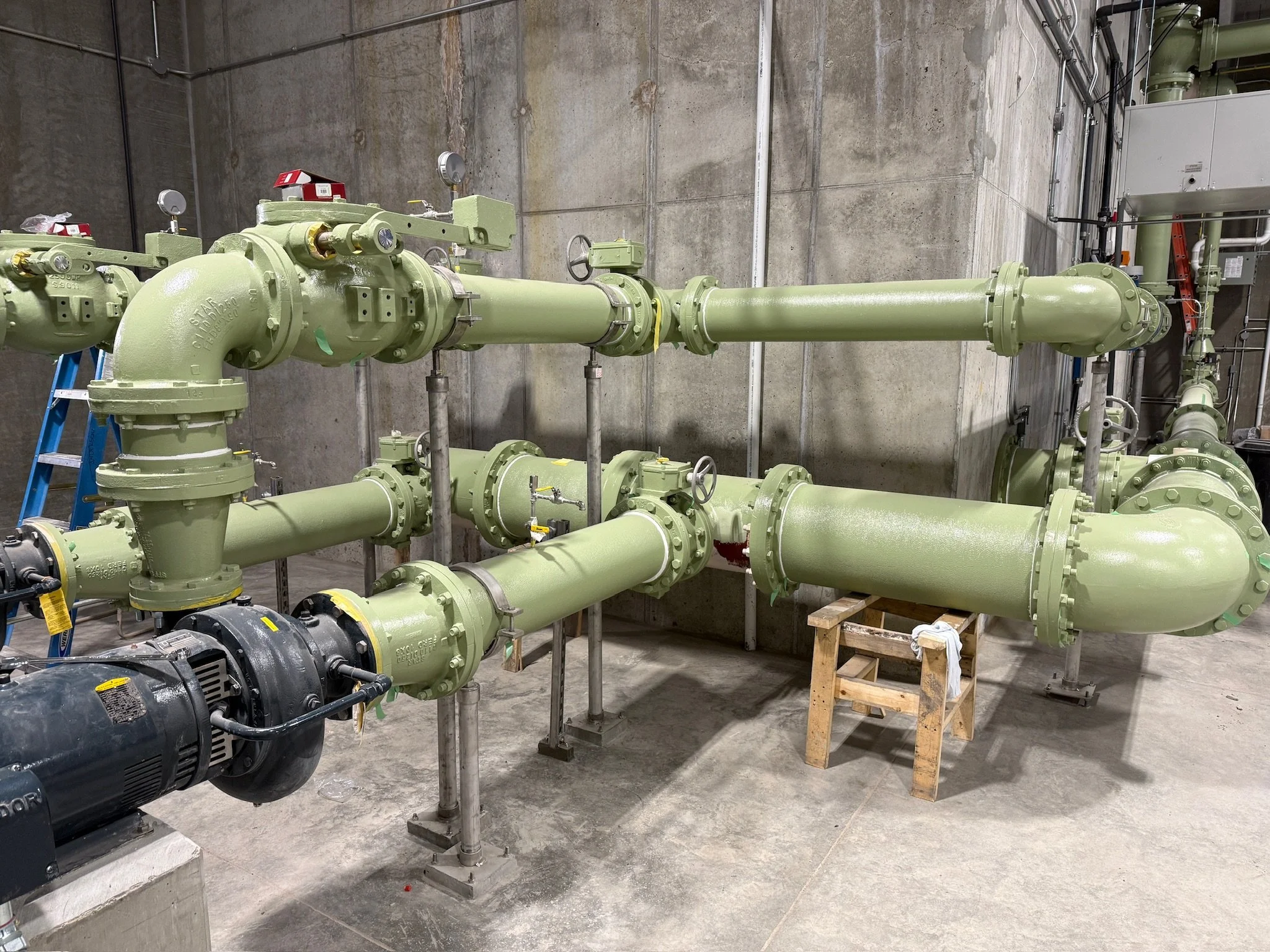 Green industrial pipes and equipment in a concrete room, supported by metal stands, with a motor or pump at the foreground left.