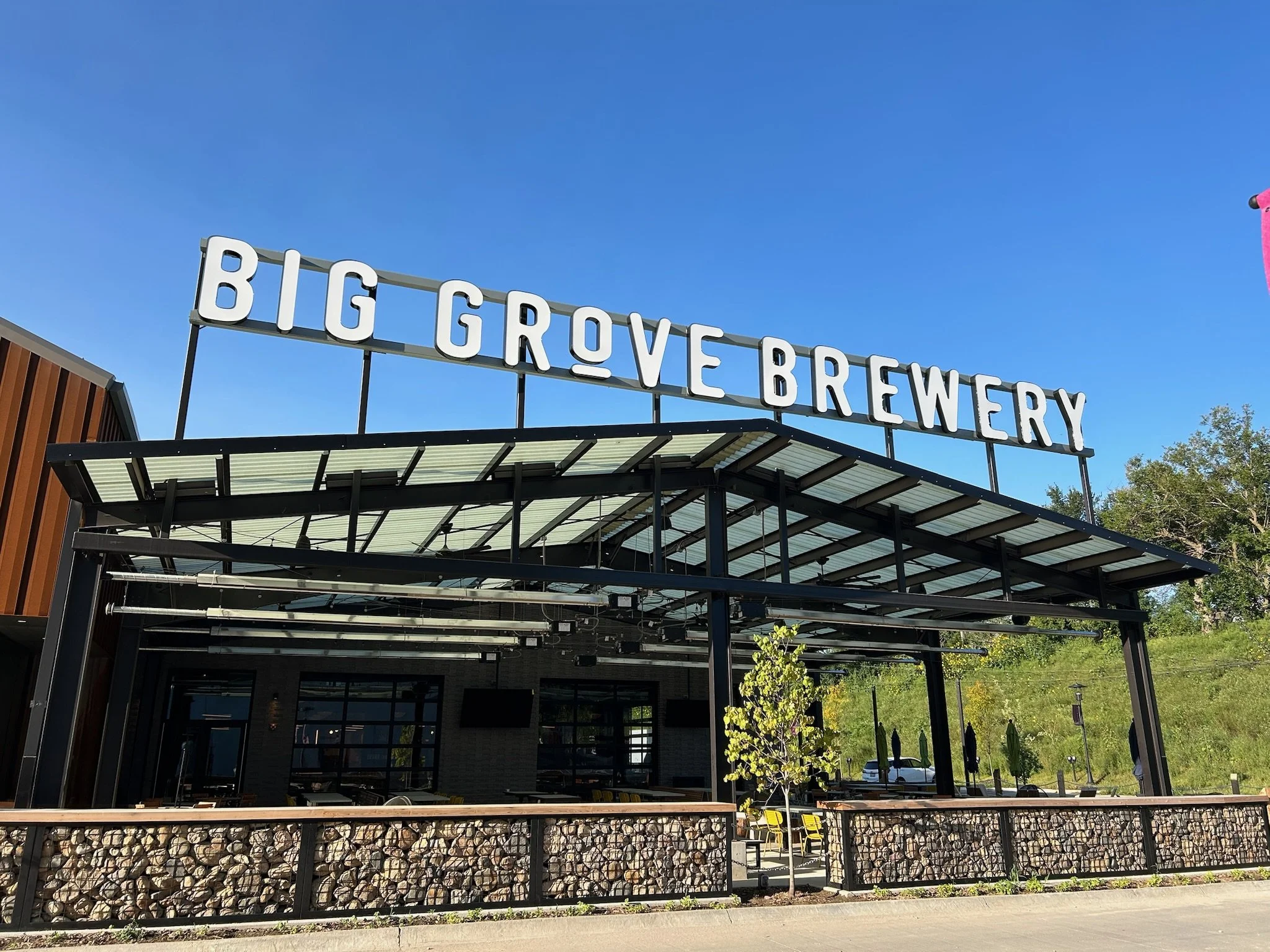 Sign for Big Grove Brewery against a clear blue sky with trees and outdoor seating area in the background.