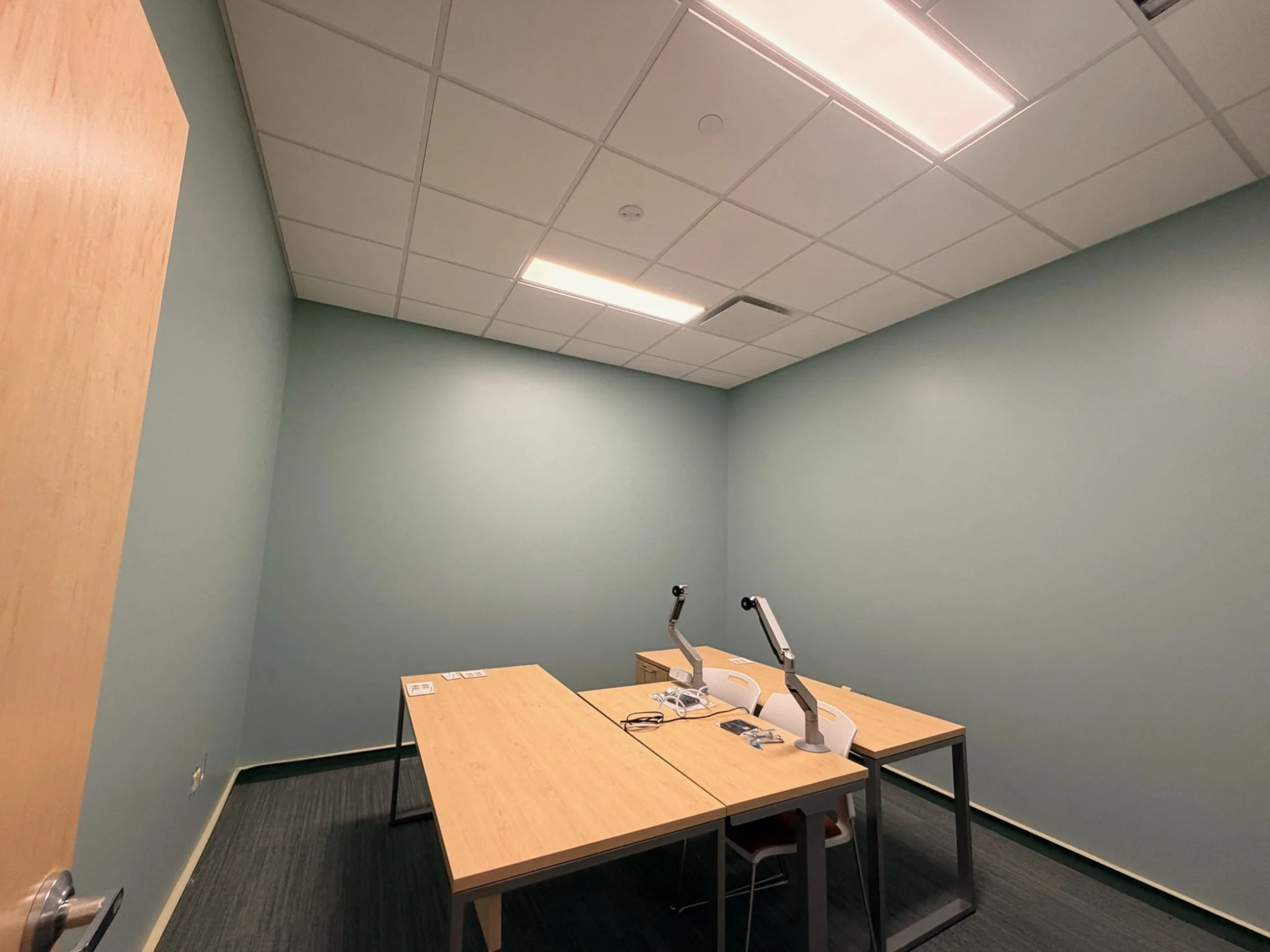 Empty meeting or training room with a light green wall, a drop ceiling with fluorescent lights, and a wooden table with chairs and some electronic equipment on it.