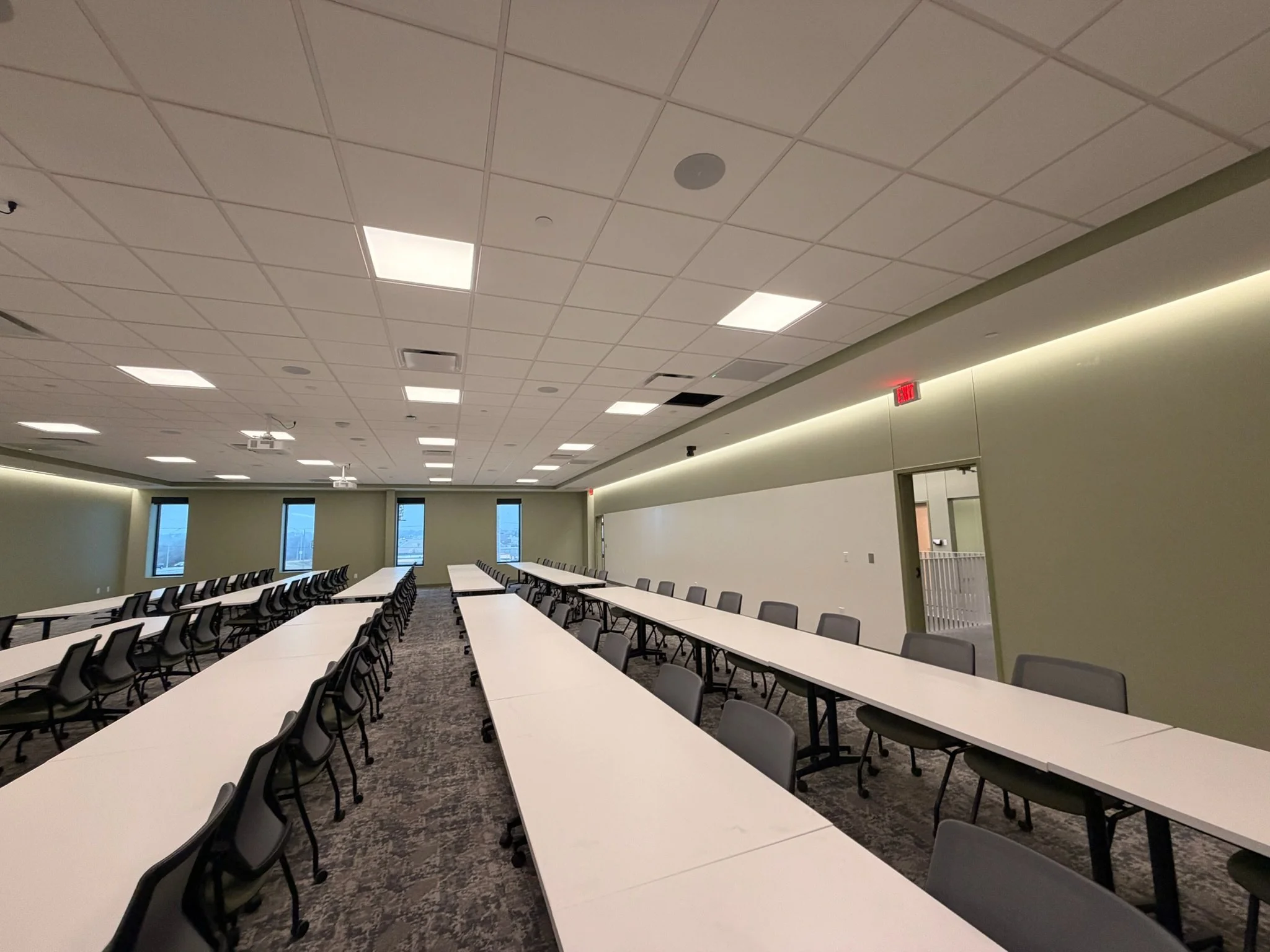 A spacious conference room with long white tables arranged in rows and black chairs around them. The ceiling has acoustic tiles with recessed lighting. The room has windows along one wall, and an exit sign is visible near the doorway.