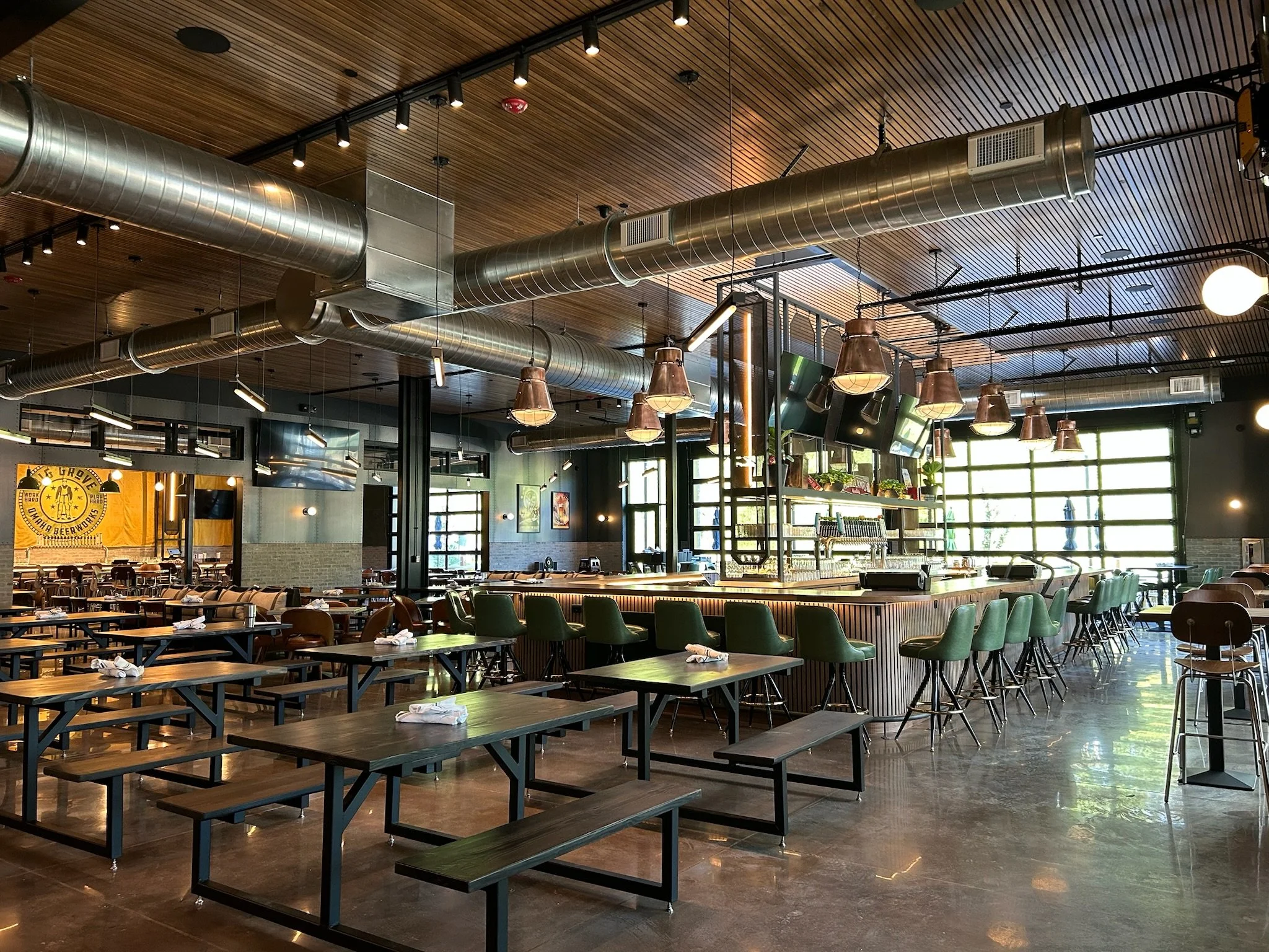 Interior of a modern restaurant or cafe with wooden ceiling, industrial ventilation ducts, hanging lights, and rows of tables and chairs.