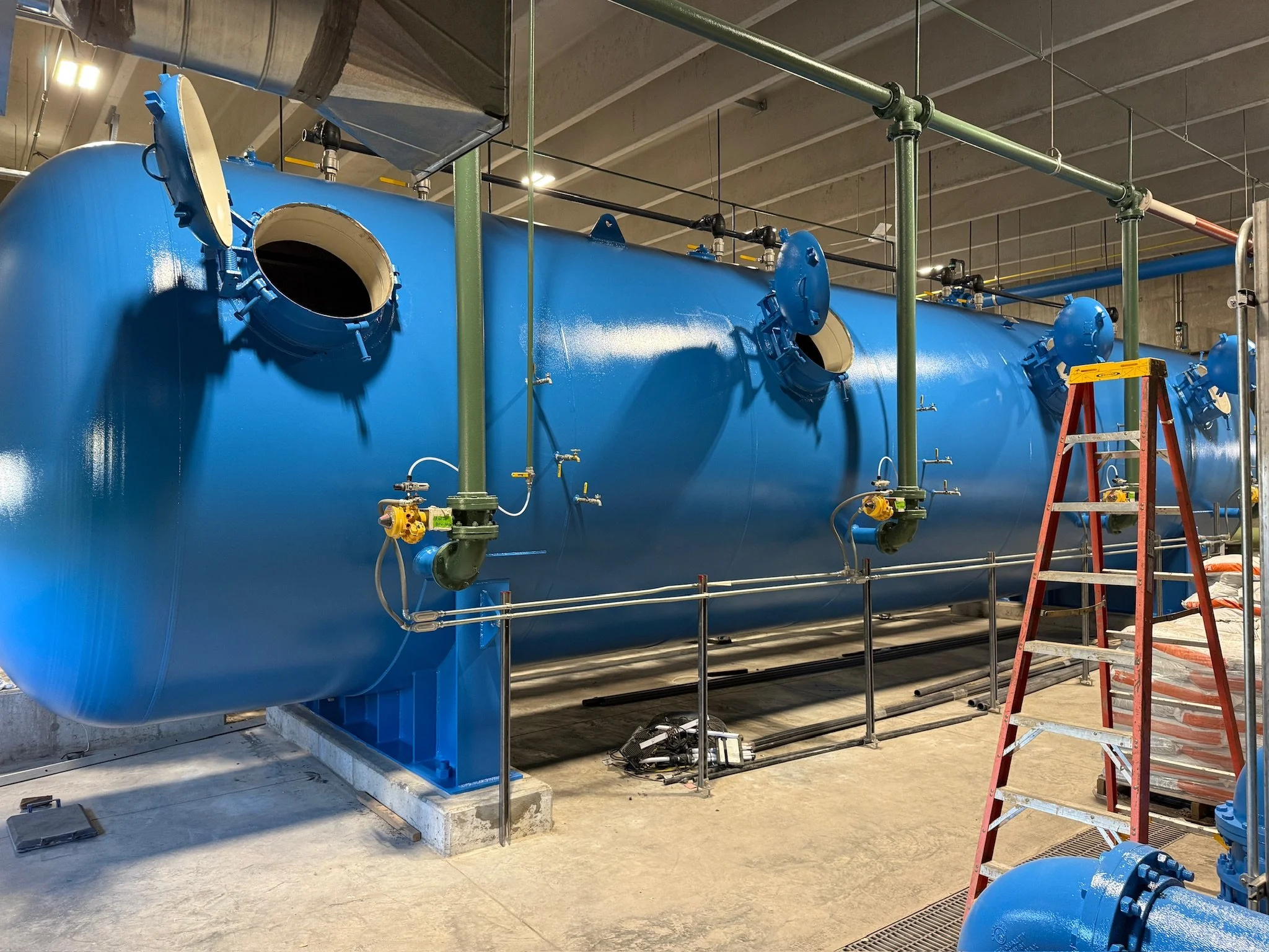 Large blue industrial tank with open manhole on a construction site, surrounded by green scaffolding and piping, with a red ladder leaning against it.