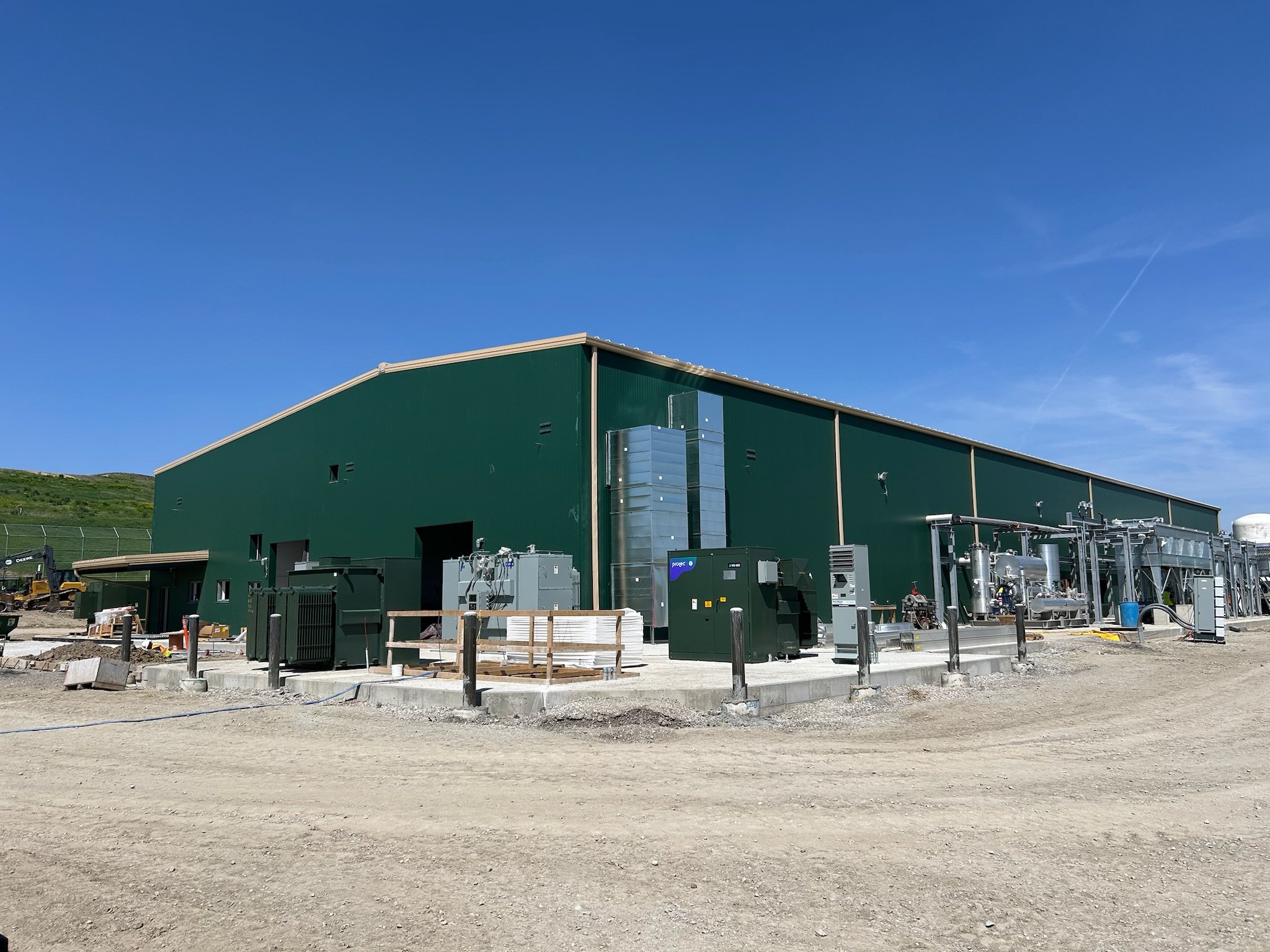 A large green industrial building under construction with various pipes, equipment, and construction materials in front. Clear blue sky above and an uneven dirt ground in the foreground.
