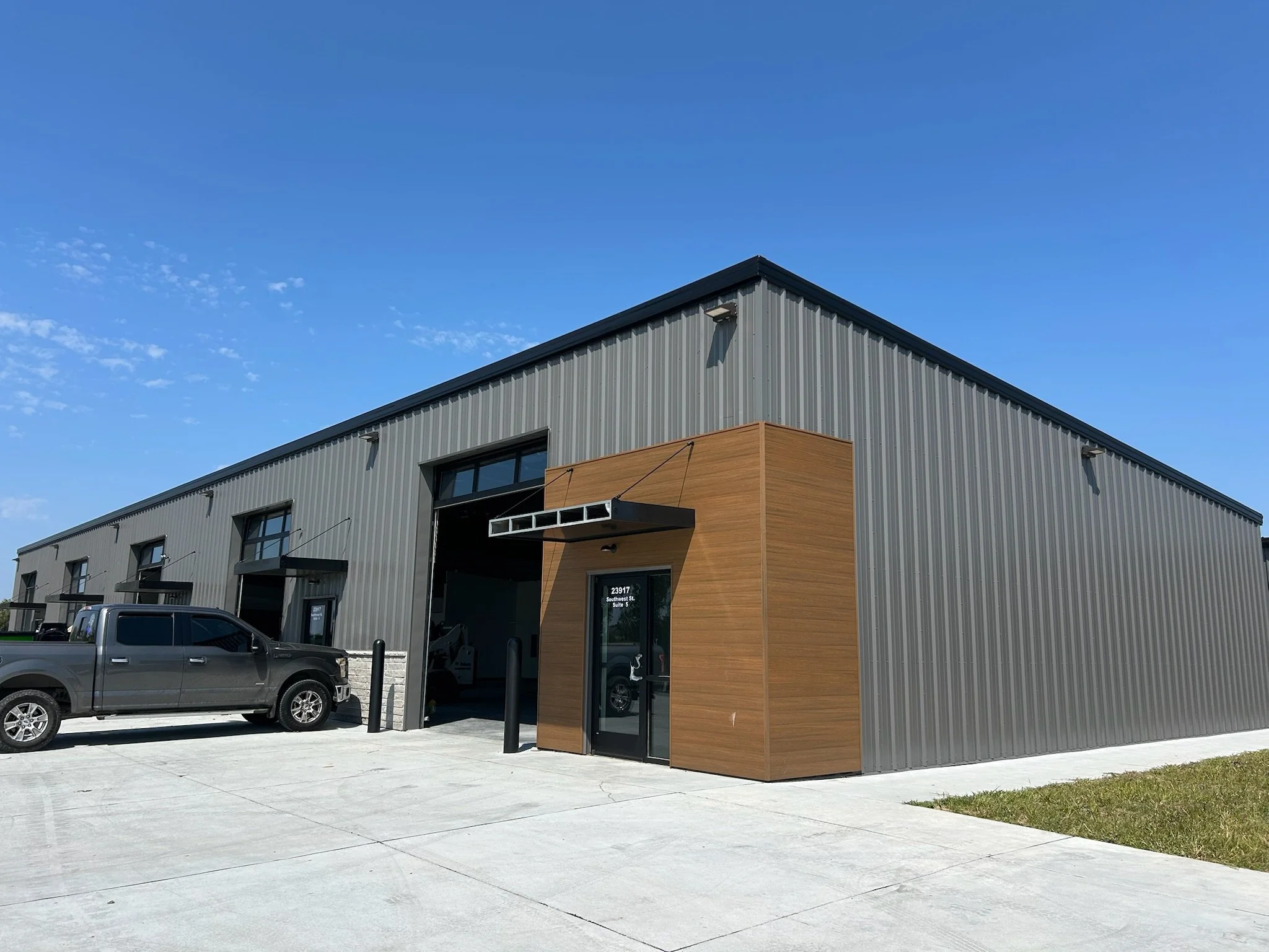 A modern commercial building with gray steel siding and a wood accent on the facade, featuring large windows and a black front door. There are black posts in front of the entrance, parked trucks nearby, and a bright blue sky overhead.