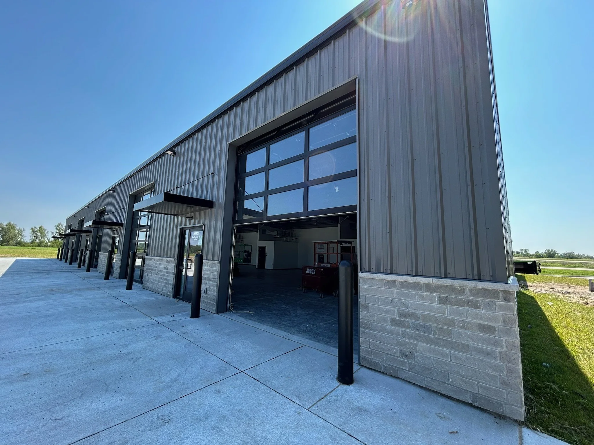 Exterior of a modern industrial building with large glass garage doors, black bollards, and a concrete sidewalk, under a clear blue sky with the sun shining.