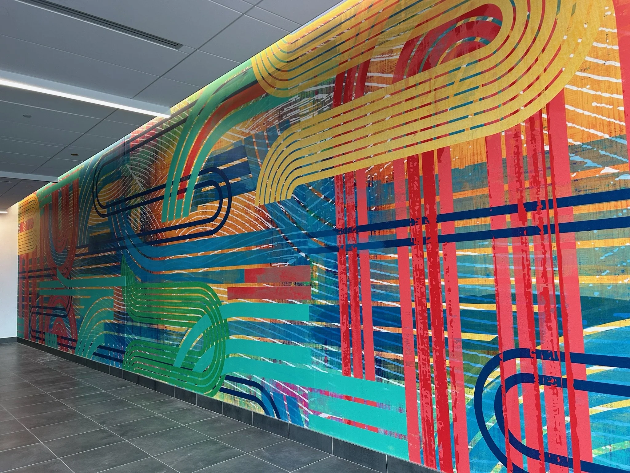 A colorful abstract mural with swirling lines and patterns in red, yellow, green, and blue, displayed on a wall in an indoor space with a tiled ceiling and dark gray tile flooring.