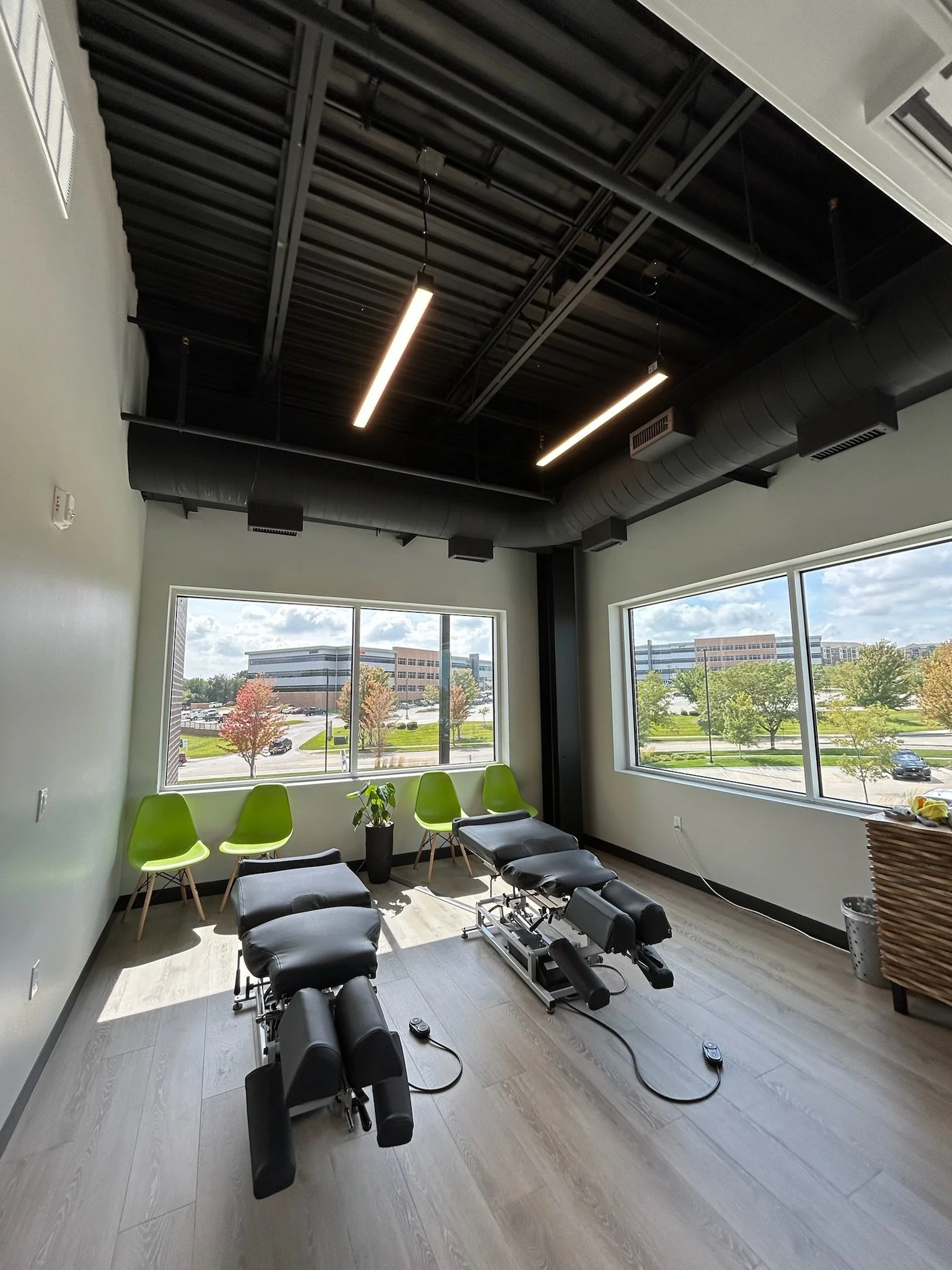 Treatment room with massage chairs, green chairs, and large windows showing trees and a parking lot outside.
