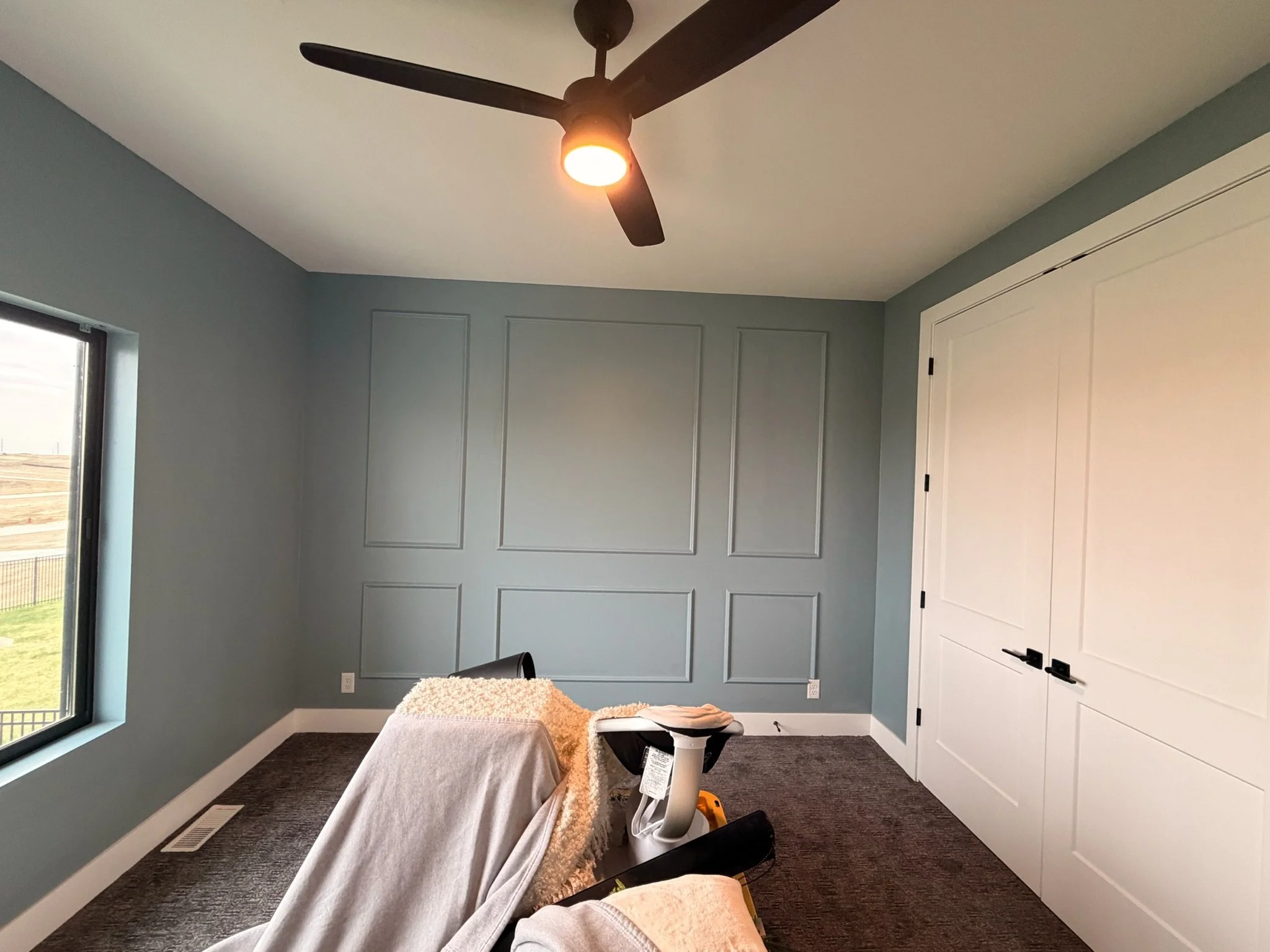 The image shows a room with blue walls, a window on the left side, a white door on the right side, and a ceiling fan with lights in the center of the ceiling. There is a table in the foreground covered with a gray cloth and some items on it, includin