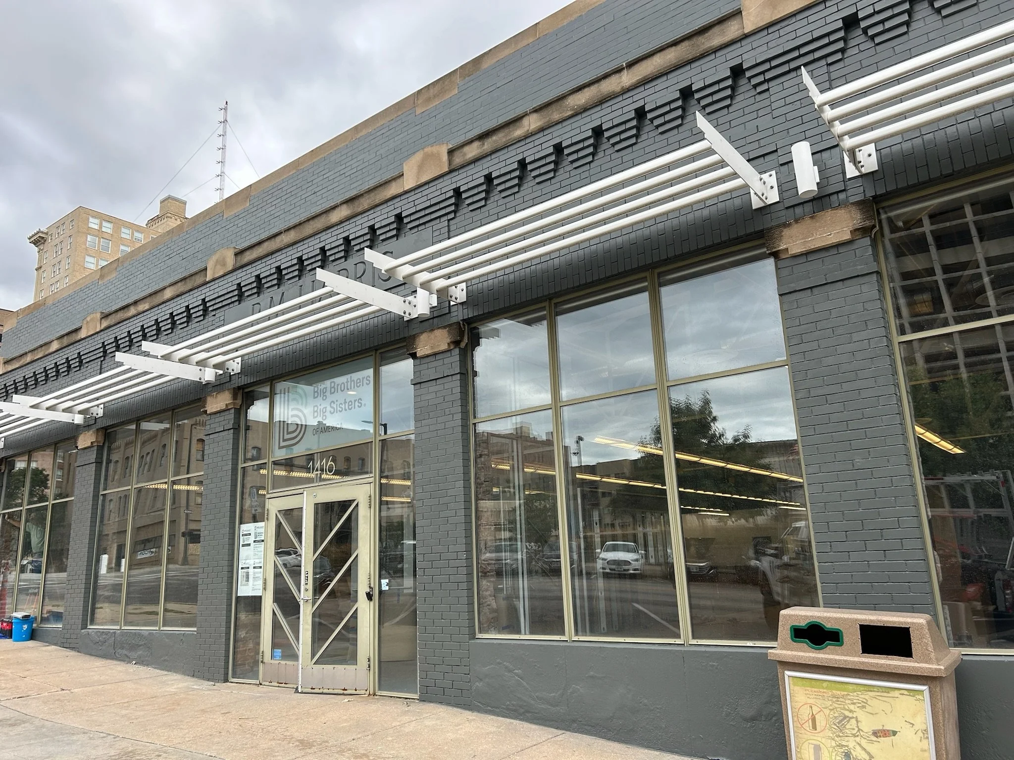 Exterior of a gray brick building with large glass windows and a white metal band pattern on the upper part. There is a sidewalk in front with a trash can and a signboard. The sky is cloudy and there are other buildings and cars reflected in the wind