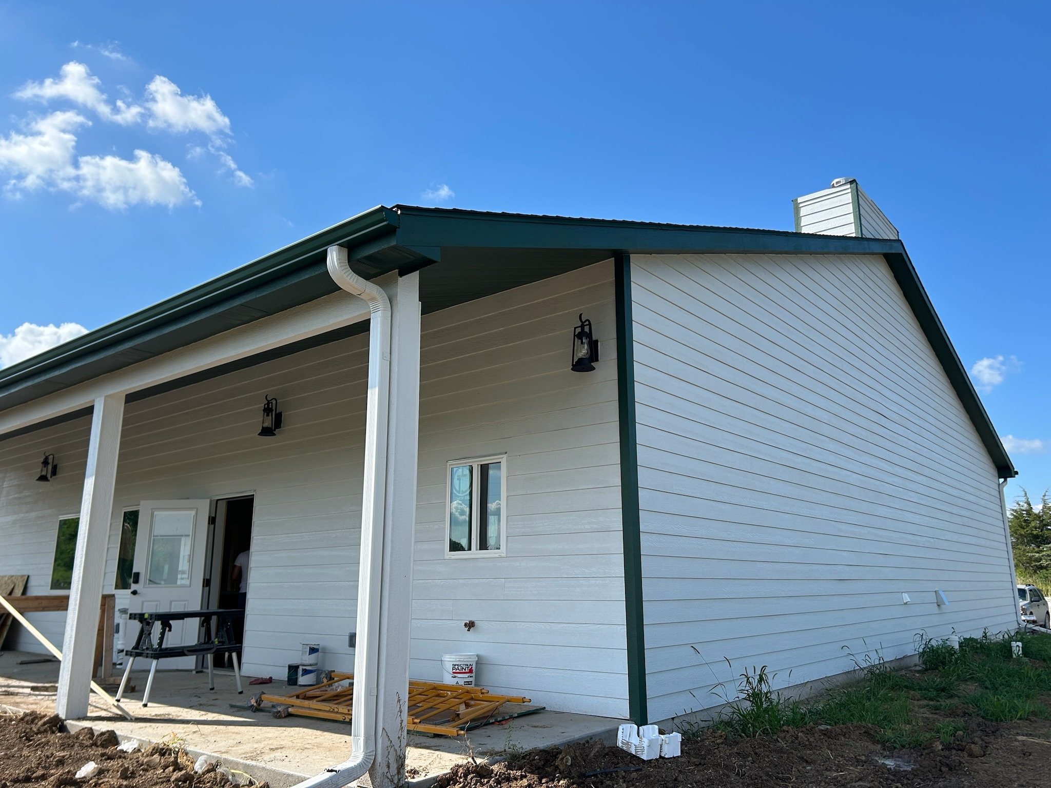Side view of a house under construction with white siding, green trim, and a porch area with supports, showing a gutter system attached to the roof and some construction tools and materials on the ground.