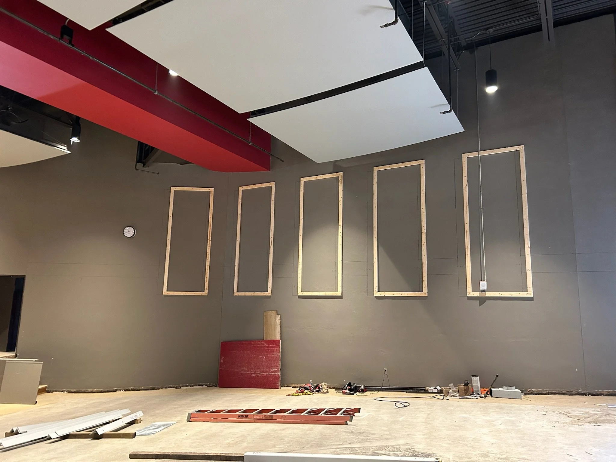 Interior of a room under construction with bare walls, a partially finished wall with wooden framing, tools and materials scattered on the floor, a clock on the wall, and ceiling panels being installed.