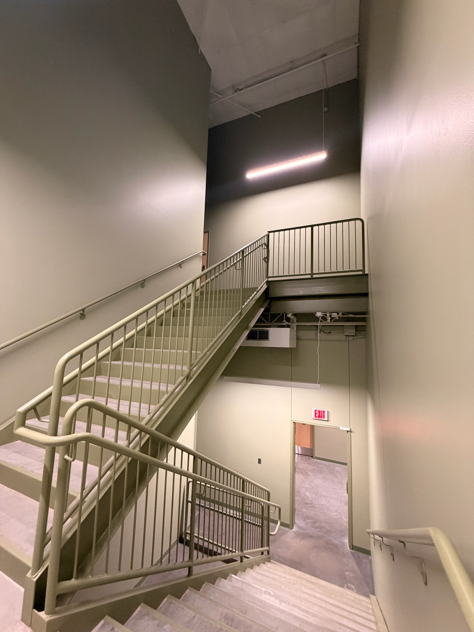 An indoor stairway with beige railings and stairs, leading to a second-floor landing. There is a gray wall on one side and a beige wall on the other, with ceiling lighting and an exit door at the bottom of the stairs.