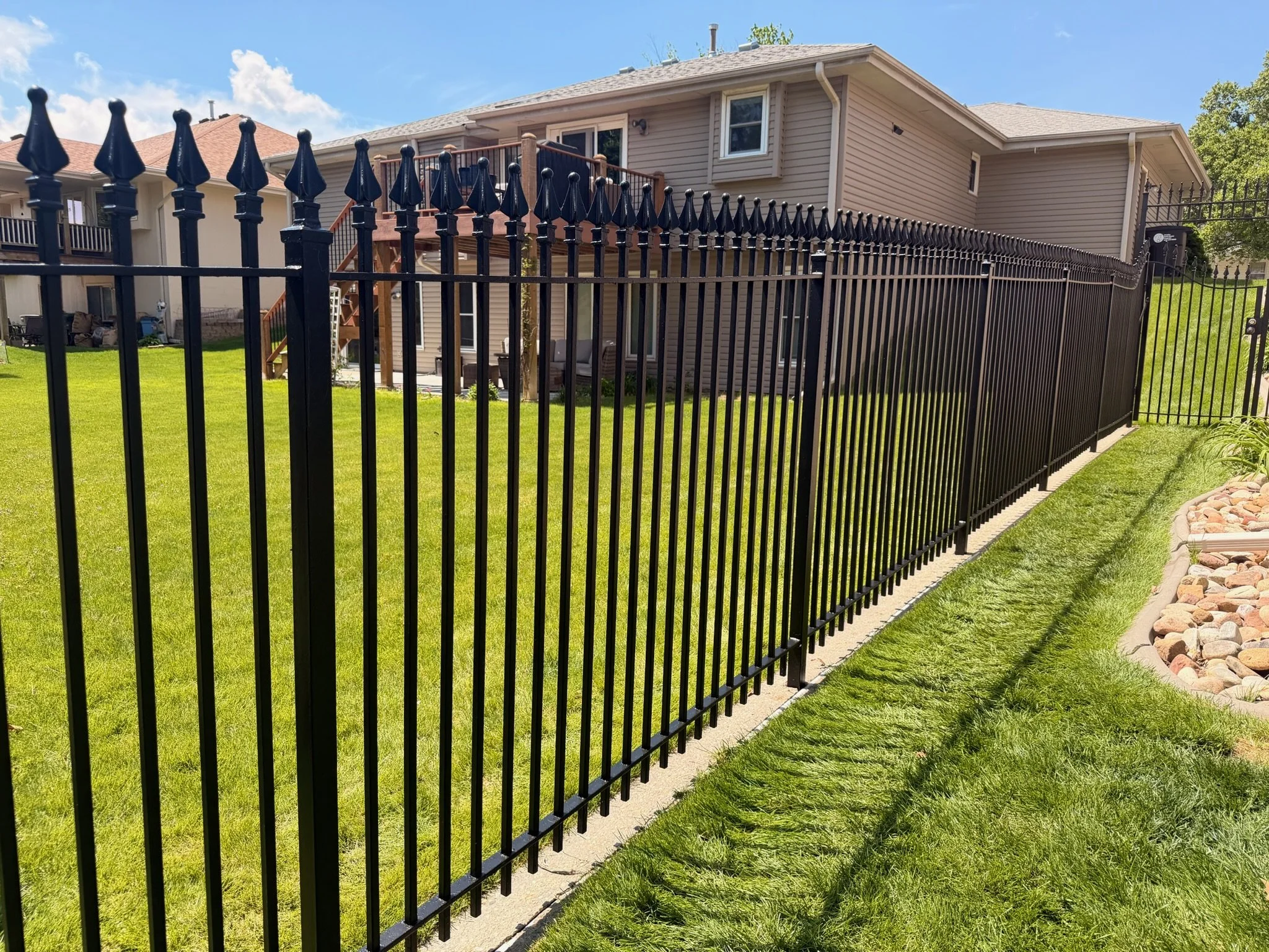 A black metal fence with pointed finials surrounds a green lawn in the backyard of a beige house, with a wooden deck and stairs visible. The sky is blue with a few clouds.