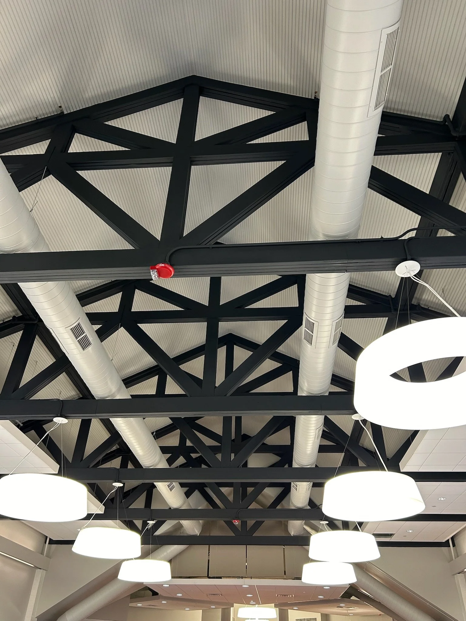 Ceiling of an indoor space featuring white circular light fixtures, black steel beams forming a geometric pattern, and large white duct pipes.