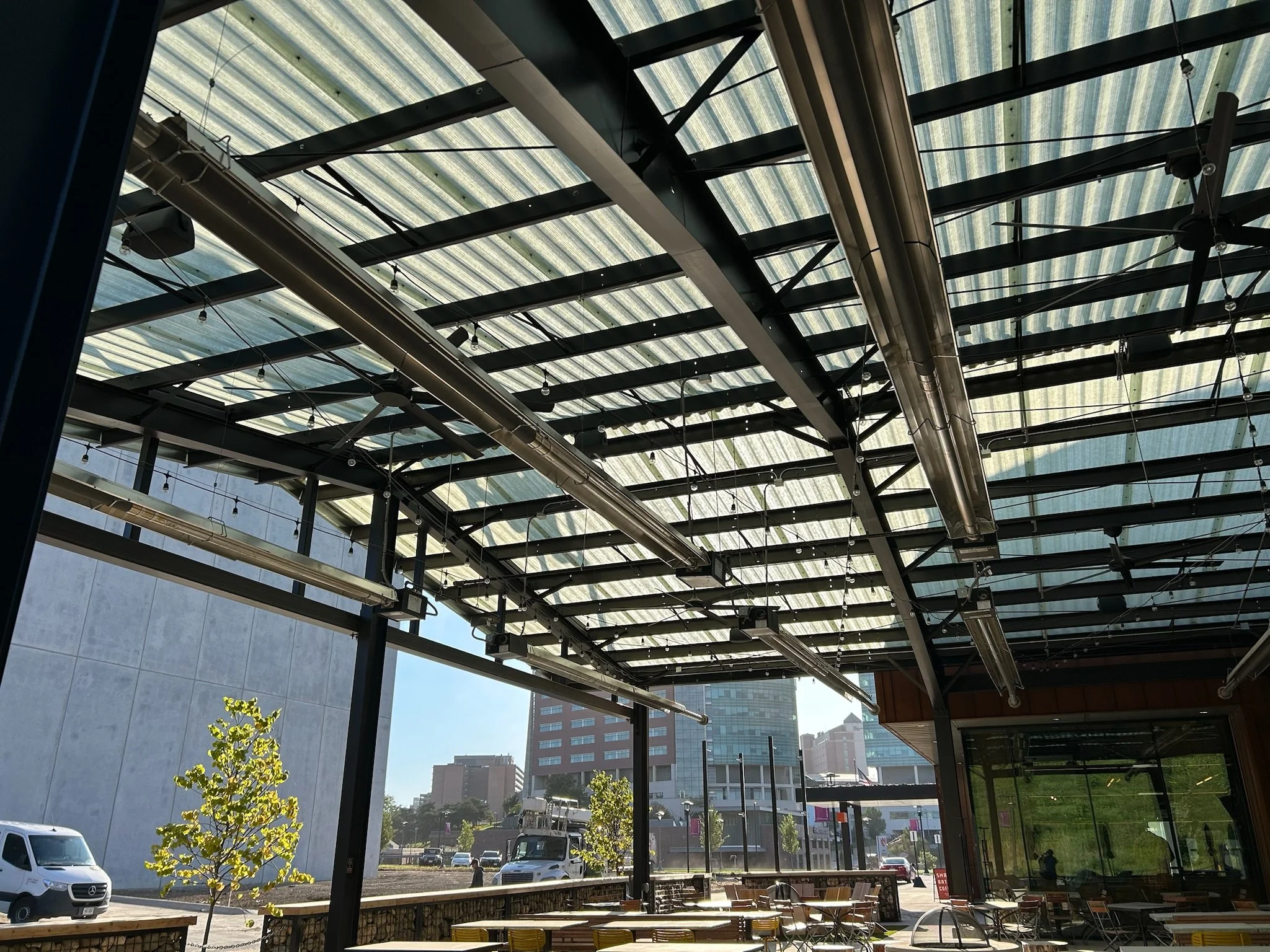 View of an indoor space with a glass roof, ceiling fans, and outdoor cityscape visible through large windows, with trees and parked cars outside.