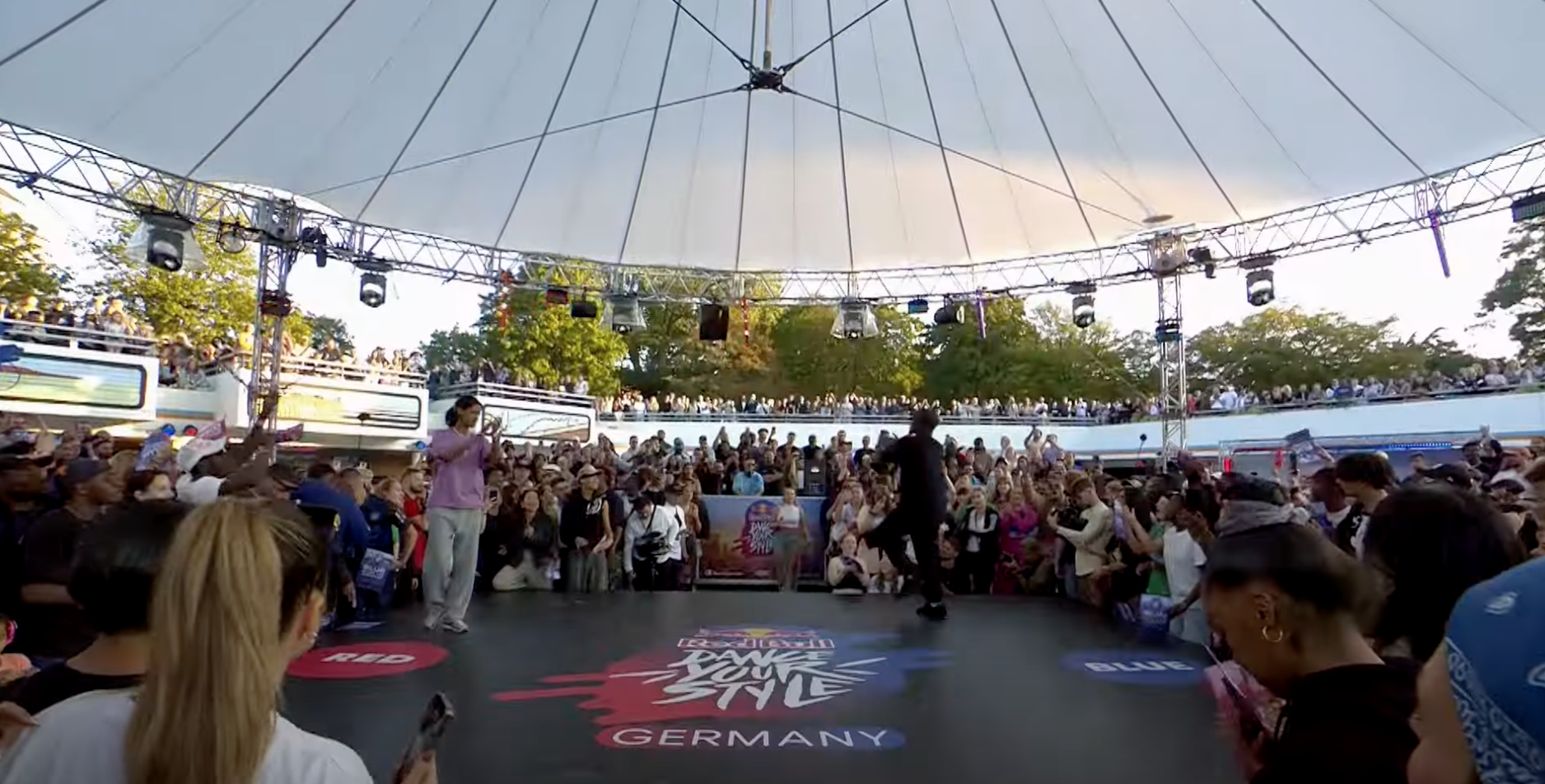 Menschenmenge bei einem Event unter einer großen schwarzen Zeltkuppel, auf einer Bühne mit dem Text 'TAKE YOU' und 'GERMANY'