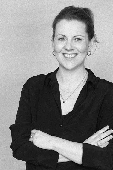 Eine Frau mit braunen Haaren, die lächelnd in die Kamera schaut, trägt eine schwarze Bluse und steht vor einem neutralen Hintergrund.