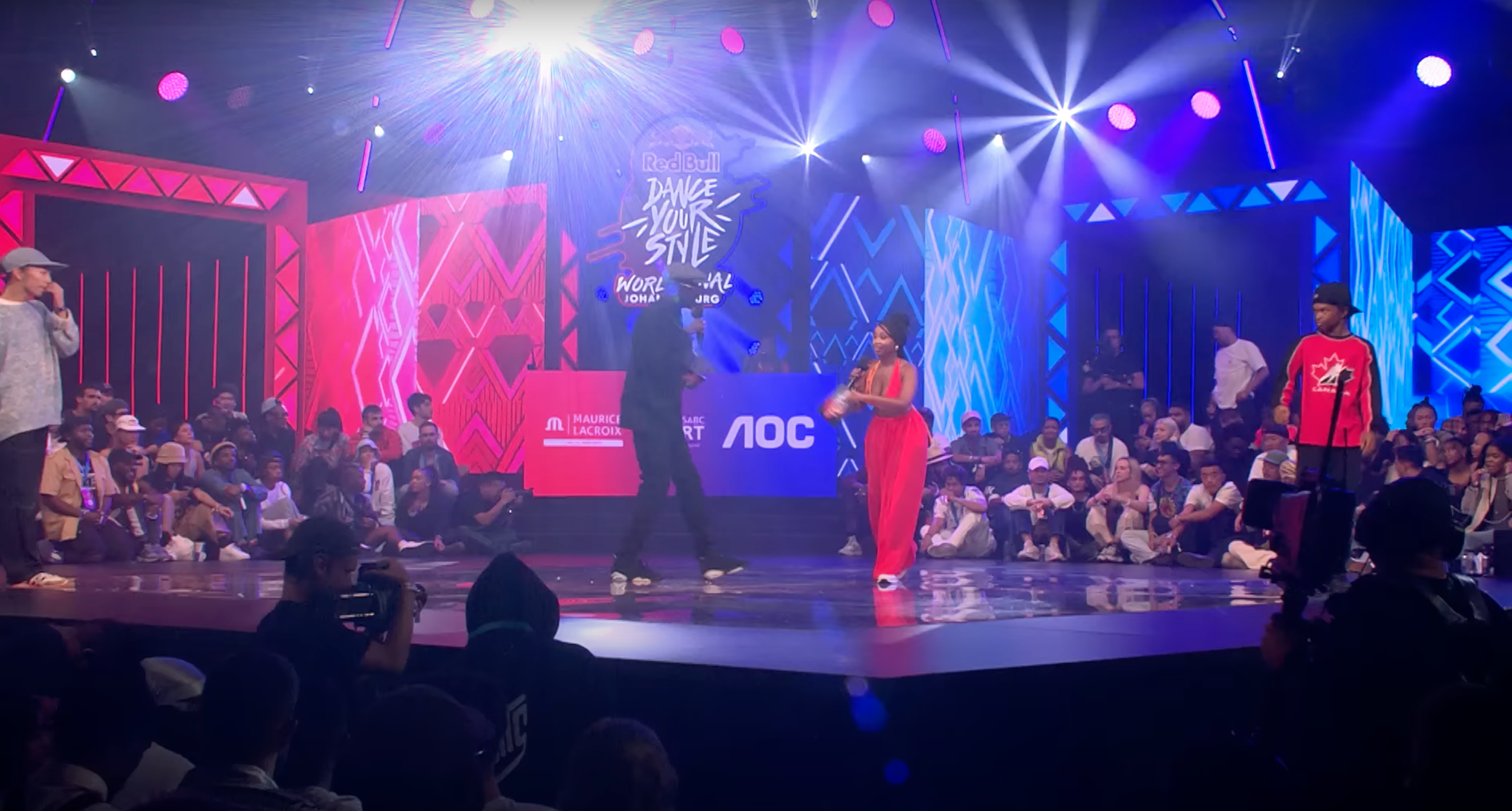 Tanzwettbewerb auf einer Bühne mit Live-Publikum und bunten Lichteffekten, hinter einem DJ-Pult mit Neon-Schriftzug 'Red Bull Dance Your Style'.