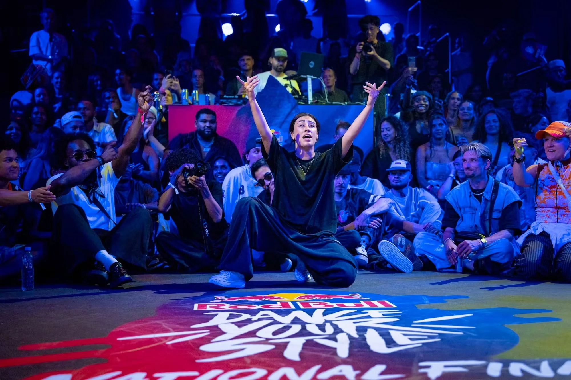 Eine Gruppe von Menschen sitzt und steht um einen tanzenden Menschen auf einer Bühne beim Red Bull Dance Your Style Wettbewerb, mit buntem Licht und Publikum im Hintergrund.