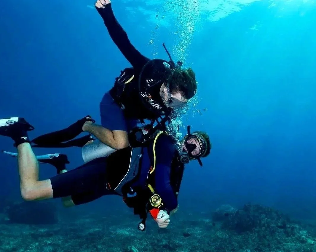 Two scuba divers underwater, one tank surfing the other, with coral reef visible below. Both divers visibly excited.