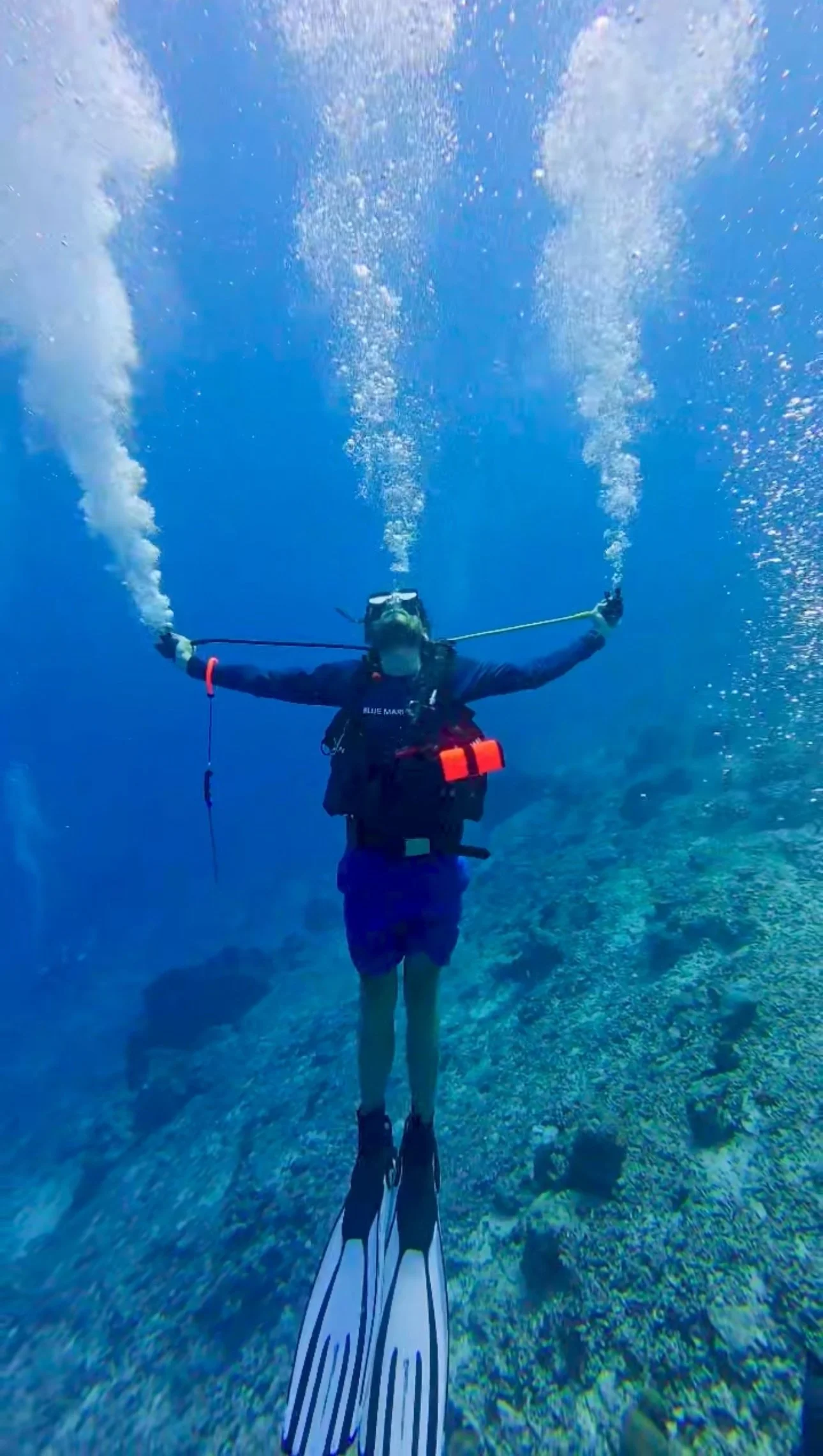 Scuba diver underwater with arms extended, surrounded by blue water and bubbles, wearing a wetsuit, mask, and fins.