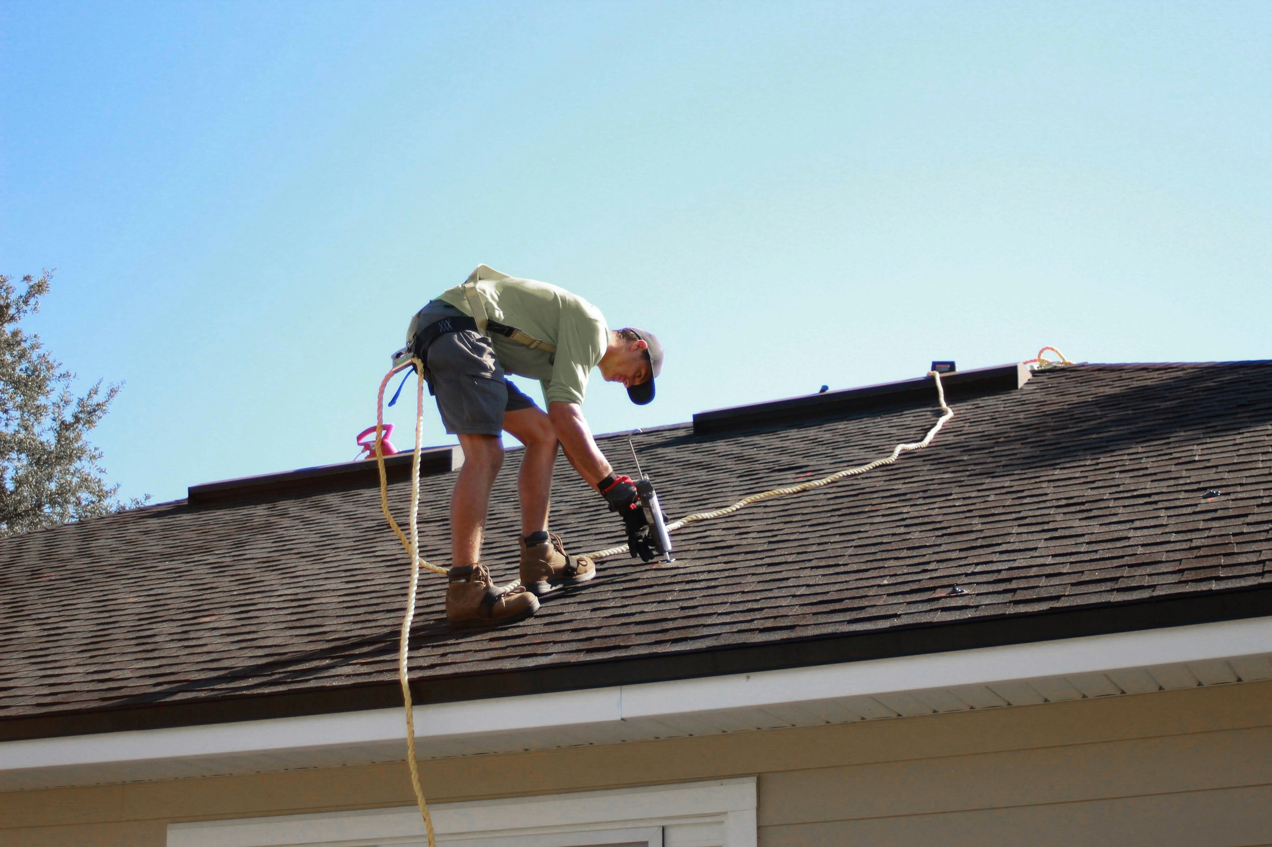 A man working on the roof of a house, using power tools and safety ropes, under a clear blue sky.