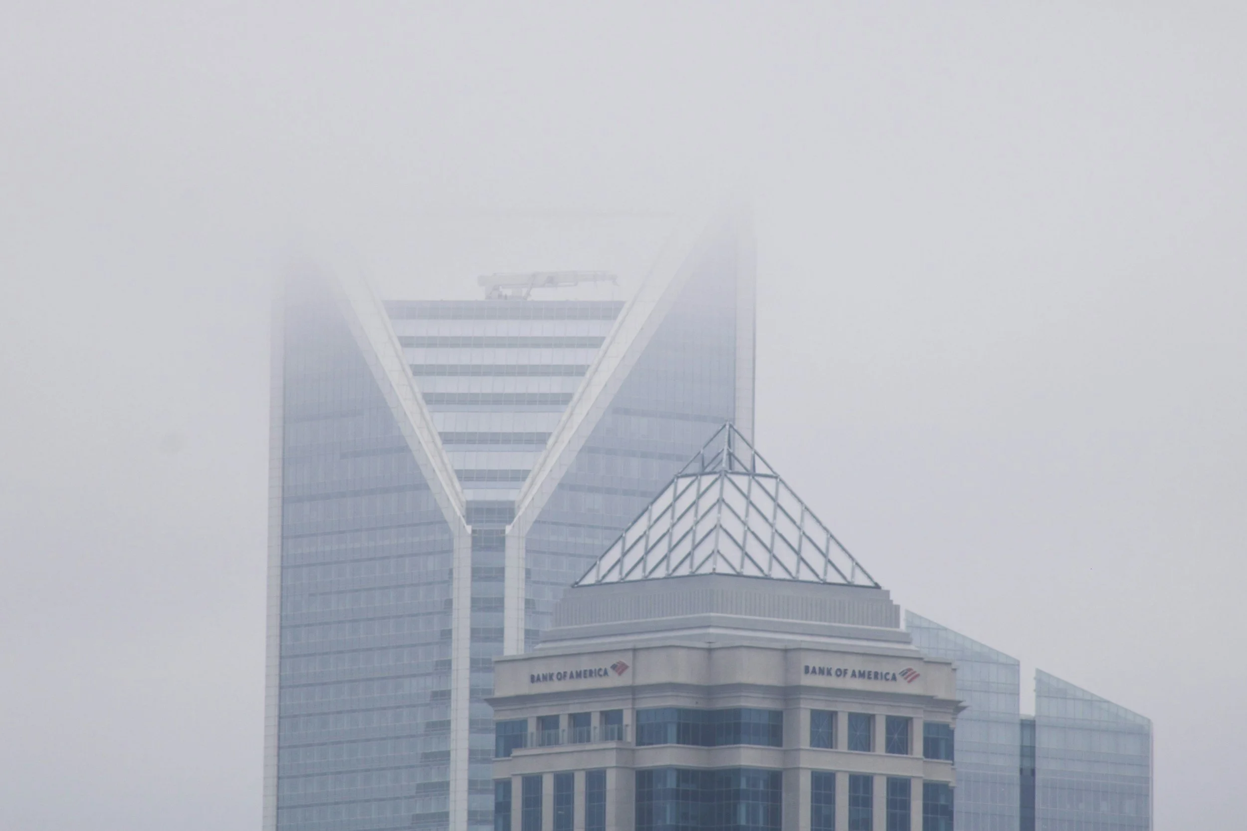 Tall modern skyscrapers in Charlotte, NC with glass facades shrouded in fog, including a building with a pyramid-shaped roof and the 'Bank of America' logo.