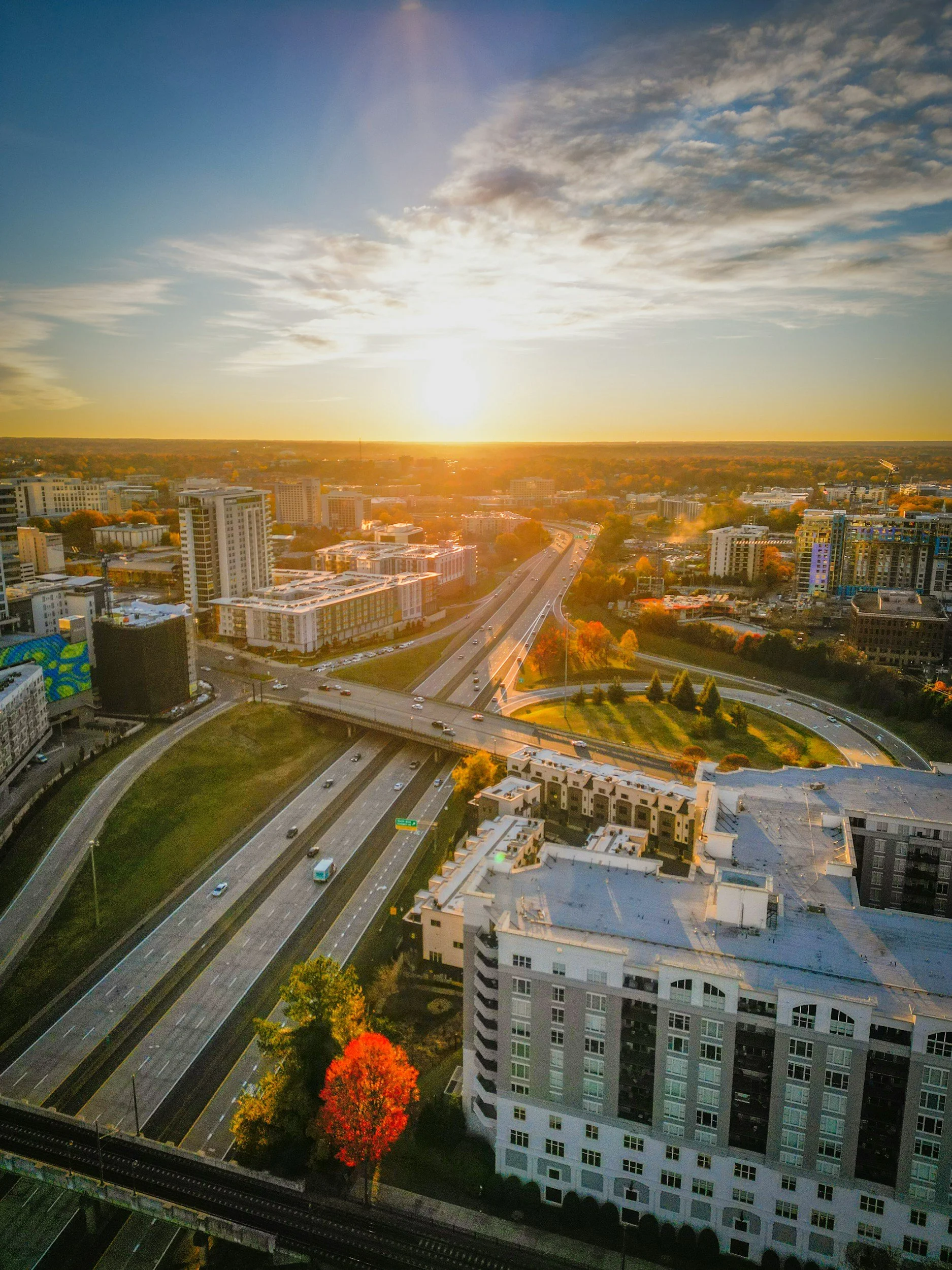 Sunset over Charlotte, NC with highways, buildings, and trees, showing a bright sky with scattered clouds.
