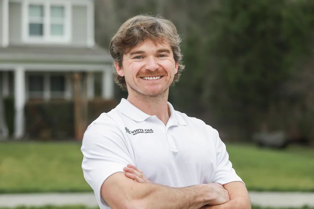 Cameron Carroll smiling with wavy brown hair wearing a white polo shirt with a White Oak Roofing logo, standing outdoors with a house and trees in the background in Concord, NC.