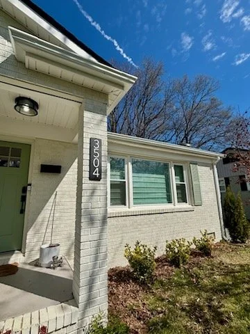 Front of a suburban house with a white brick facade, a large window, and house number 3504 on a vertical sign. Blue sky with a visible contrail behind leafless trees.
