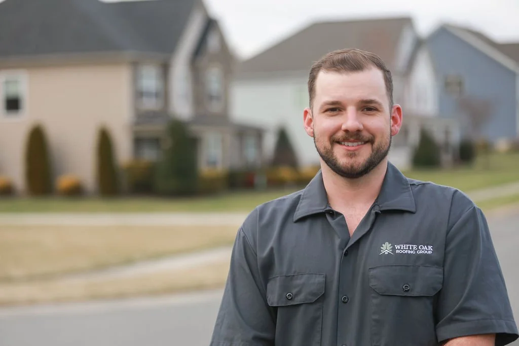Jesse Fry "The Roofing Guy" smiling in a gray White Oak Roofing work shirt with a logo on the chest, standing outdoors in a suburban neighborhood in Charlotte, NC with houses in the background.