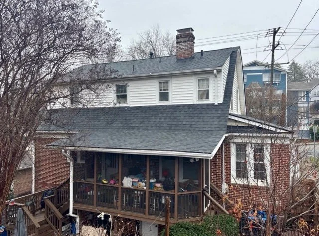 A house with a brick and white siding exterior, a screened porch with items inside, and a steep roof with a chimney. There are trees and power lines nearby.