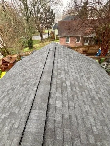 Looking down the roof of a house with gray asphalt shingles, showing the peak and surrounding trees and neighboring houses in the background.