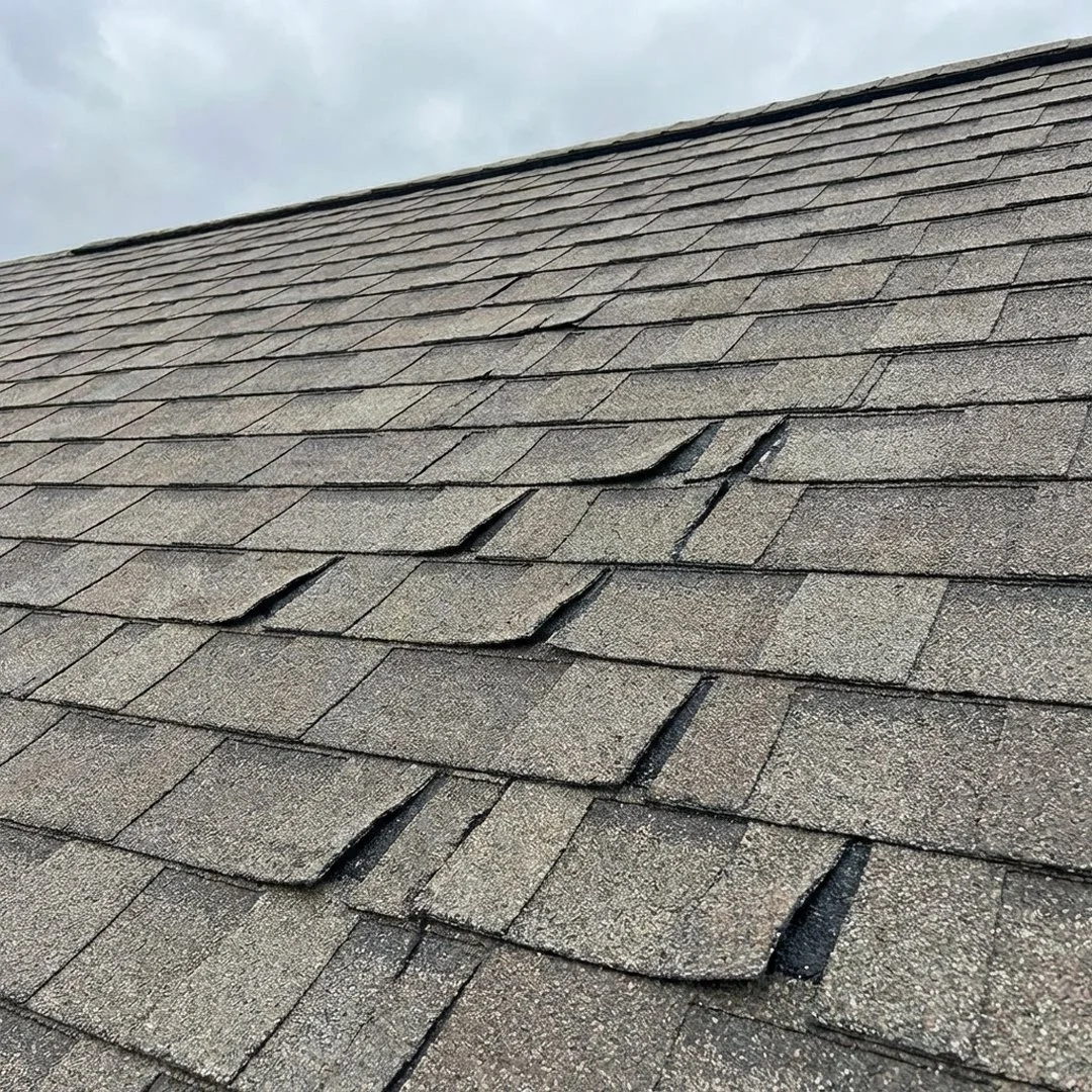 Close-up of a shingled roof with some shingles curled or damaged, under a cloudy sky.