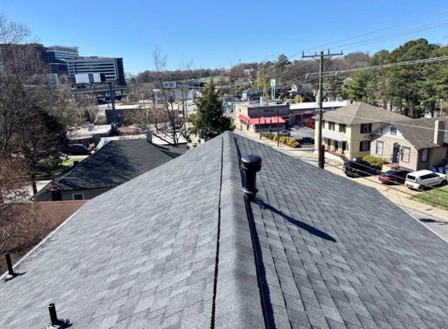 View of a rooftop with a vent pipe and rain gutter, overlooking neighboring houses and a commercial area with trees and power lines.