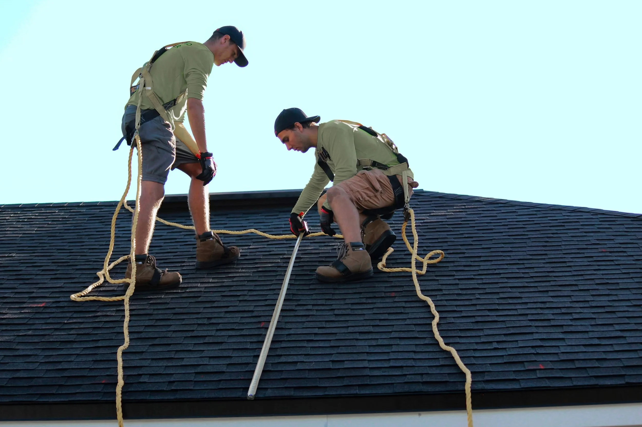 Two men working on roofing, wearing safety harnesses, using tools, on a sloped dark shingled roof during daytime.