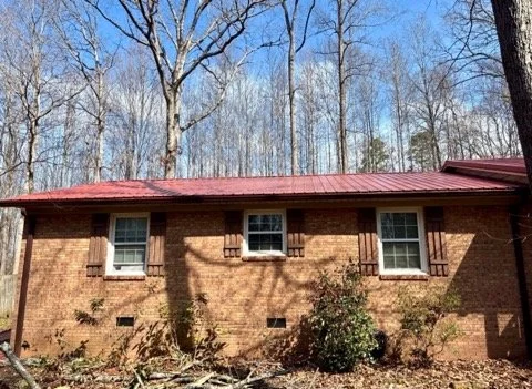 Front view of a brick house with three windows, wooden shutters, and a red metal roof, surrounded by leafless trees on a clear day.