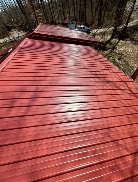 Red metal roof with sunlight and shadows, surrounded by trees and a wooded area with a shed and cars in the background.