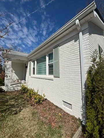 Exterior of a white brick house with a small front yard, a window with green shutters, and a bright blue sky with clouds.